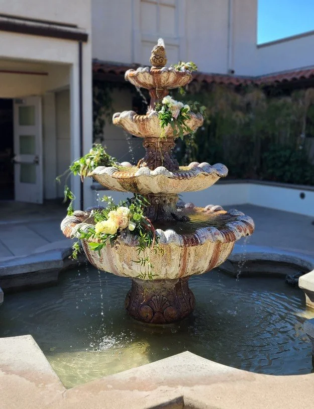 Decorative tiered outdoor fountain with flowers and greenery, set at Chaminade Resort & Spa in Santa Cruz, California. Elegant wedding flowers decorating the tiered fountain in the ceremony courtyard. 