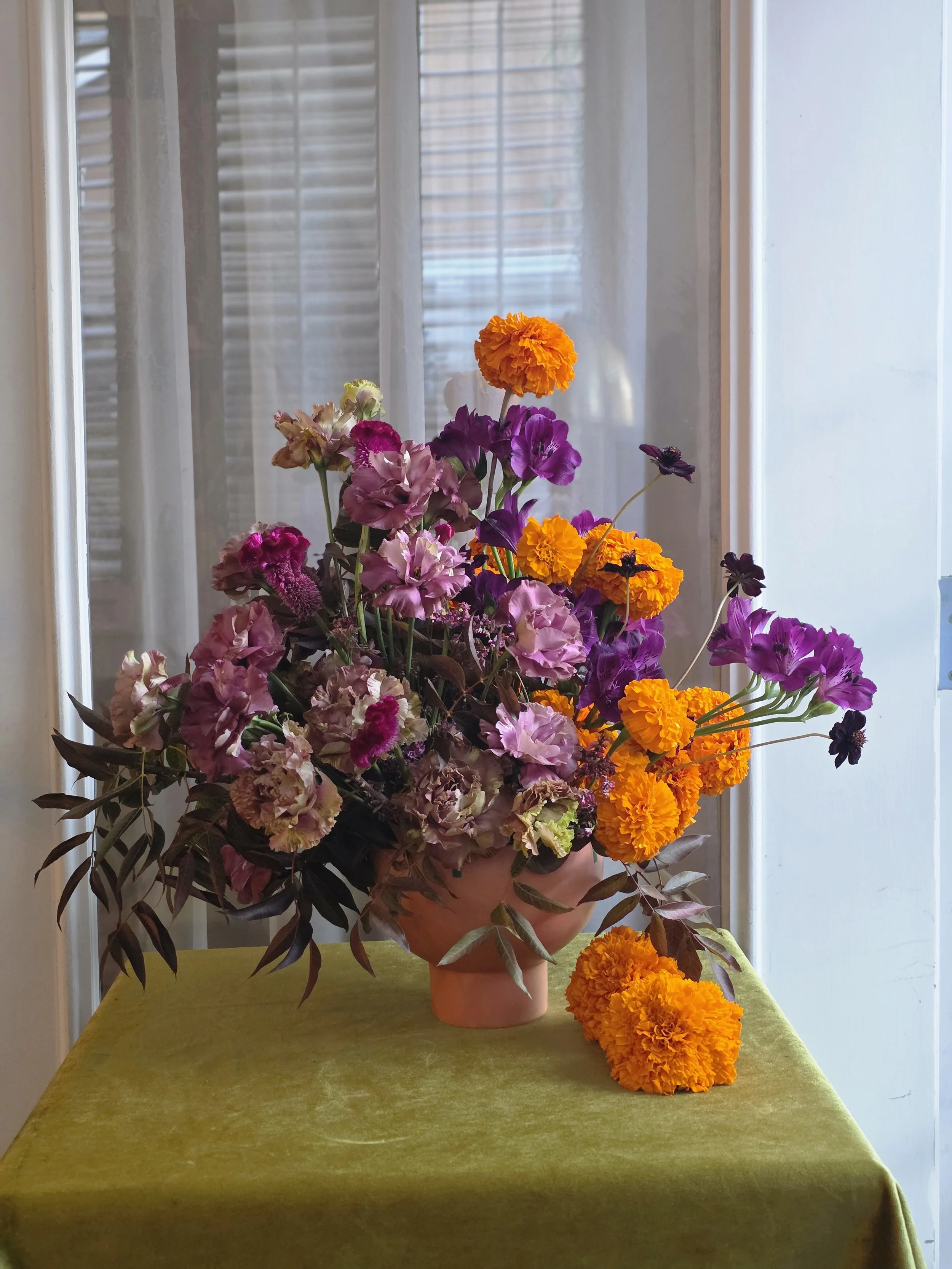 Bold pink, purple, and orange floral arrangement in a vase on a green table in natural window light, Petal and Salt Floral