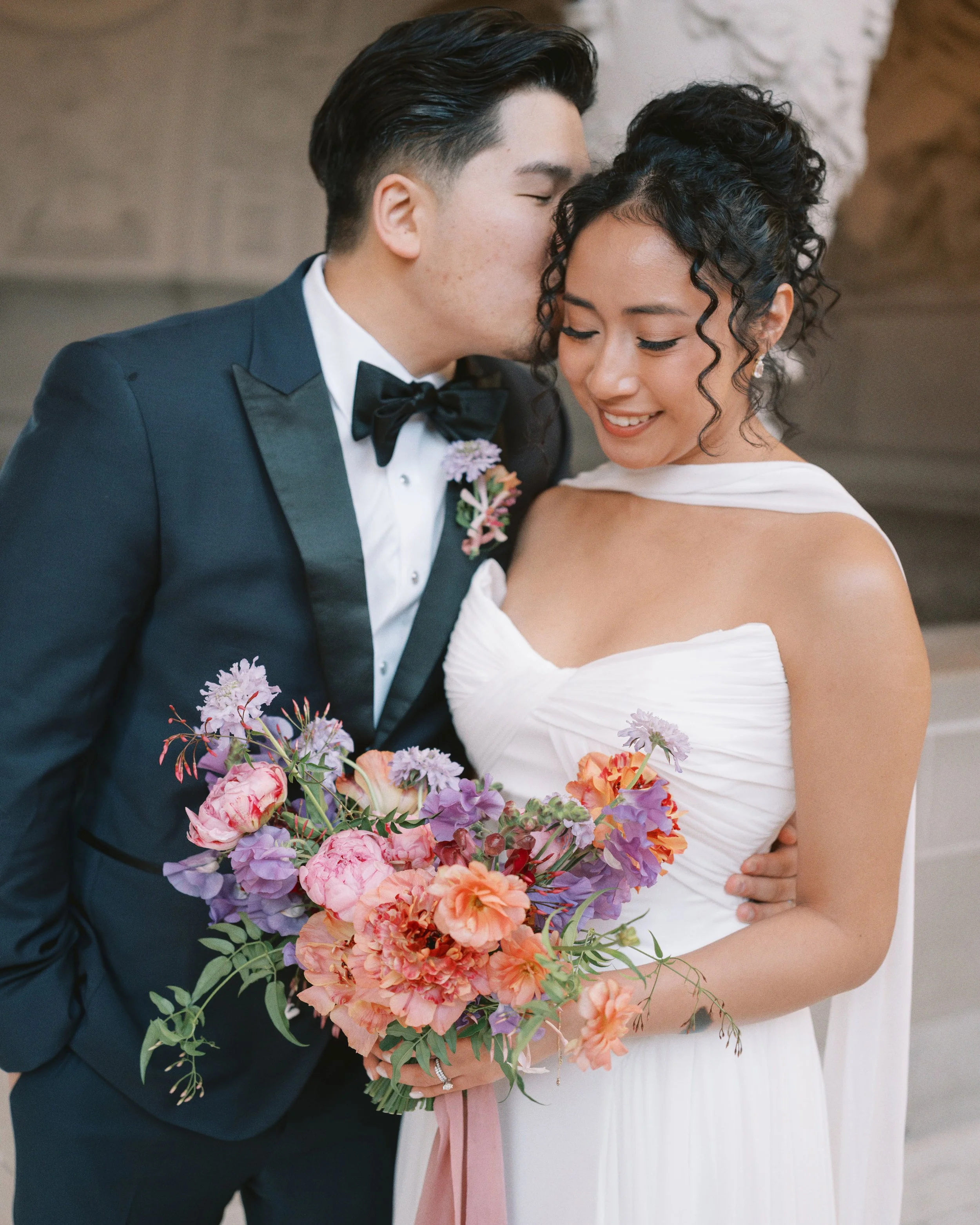 Groom kissing his bride's cheek as she smiles holding a colorful bouquet, wedding florals by Petal and Salt Floral