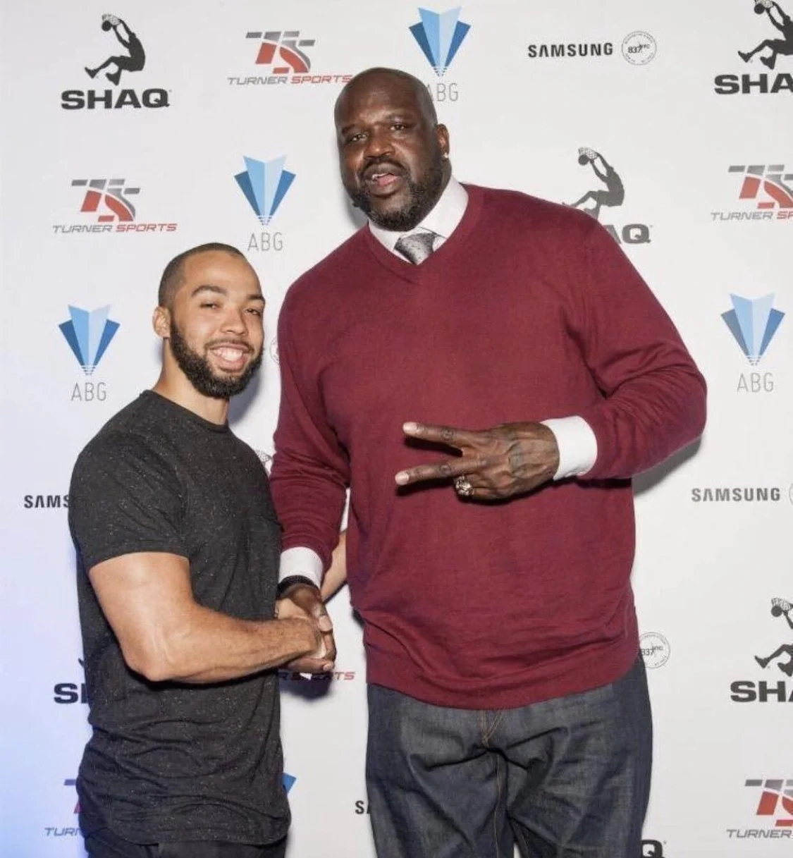 Two men in front of a step-and-repeat backdrop with logos, shaking hands. Shaquille O'Neal is on the right, taller, making a peace sign with his left hand, wearing a red sweater over a white shirt and tie. Anthony Nitti is on the left, smiling.
