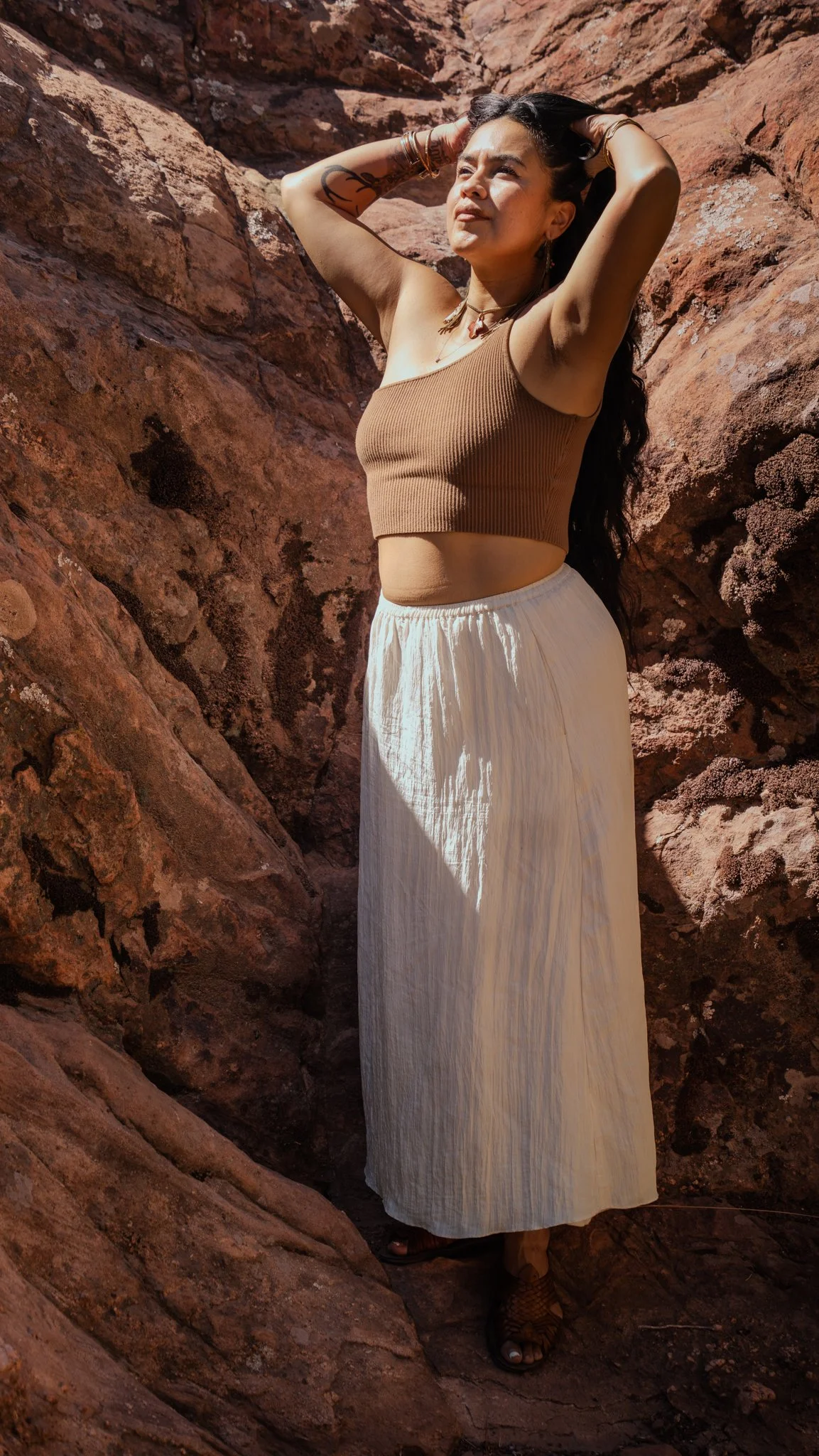 A woman with long dark hair, wearing a brown crop top and a cream-colored maxi skirt, stands amidst reddish rocks. She has her hands on her head and looks slightly upward, with sunlight shining on her face.