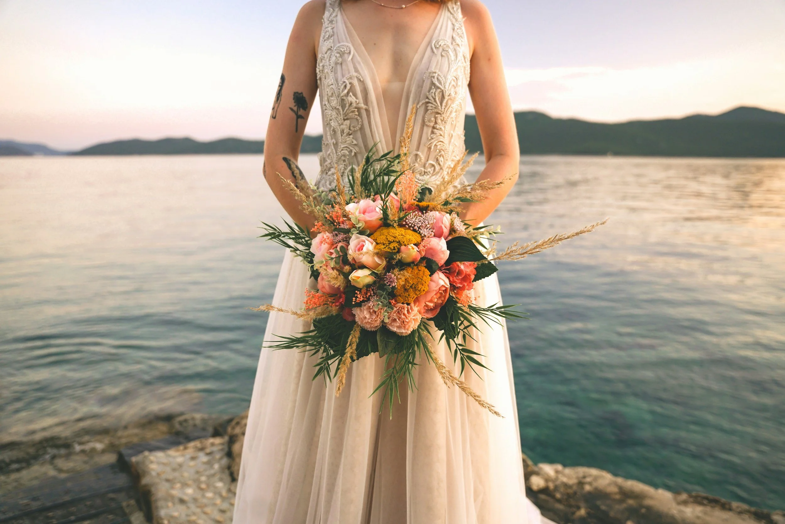 A woman in a wedding dress holding a bouquet of pink, yellow, and white flowers with green foliage, standing by a body of water with mountains in the background, during sunset.