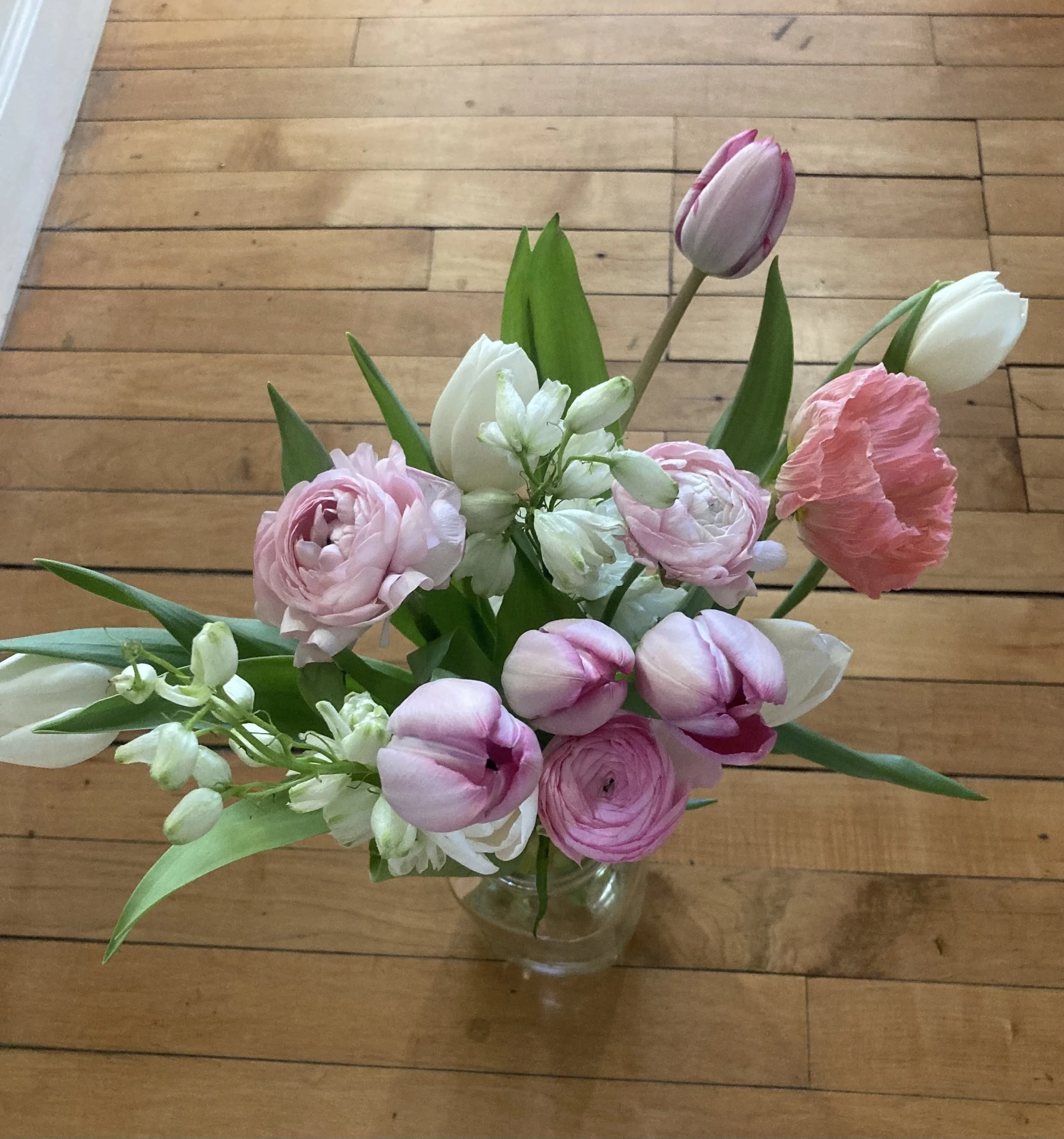 A glass vase with pink and white tulips, pink ranunculus, and white flowers, placed on a wooden floor.
