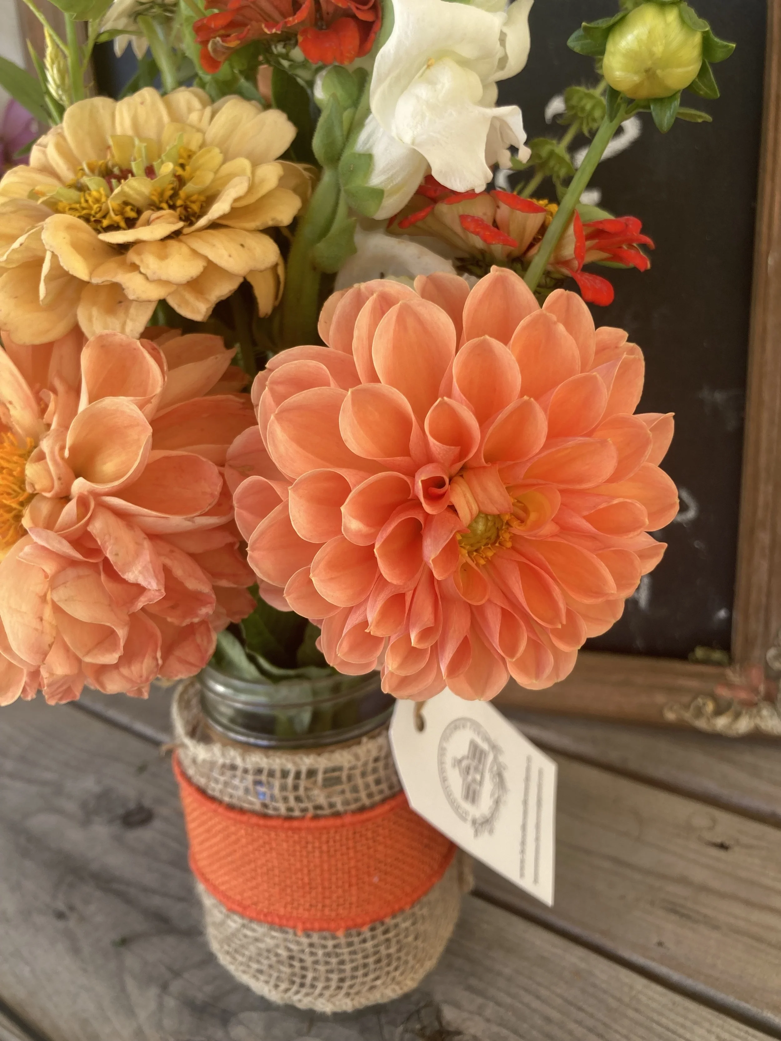 A bouquet of flowers in a glass jar, featuring a large peach-colored dahlia, yellow and cream-colored flowers with some red accents, and green buds, placed on a wooden surface.