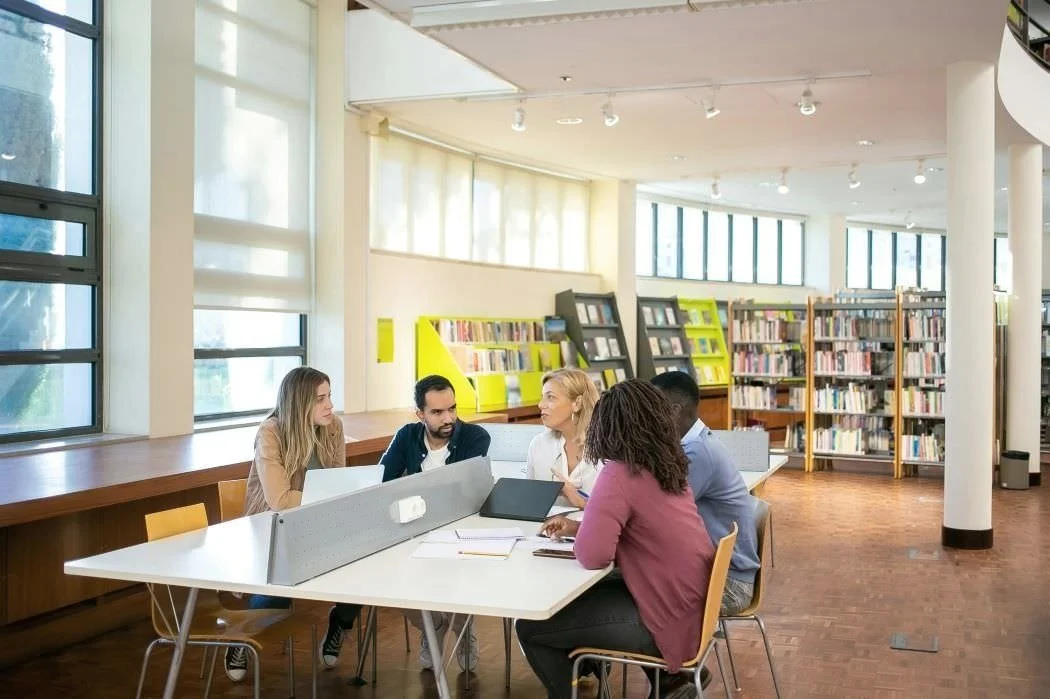 students with notebooks listen to teacher while sitting around library table