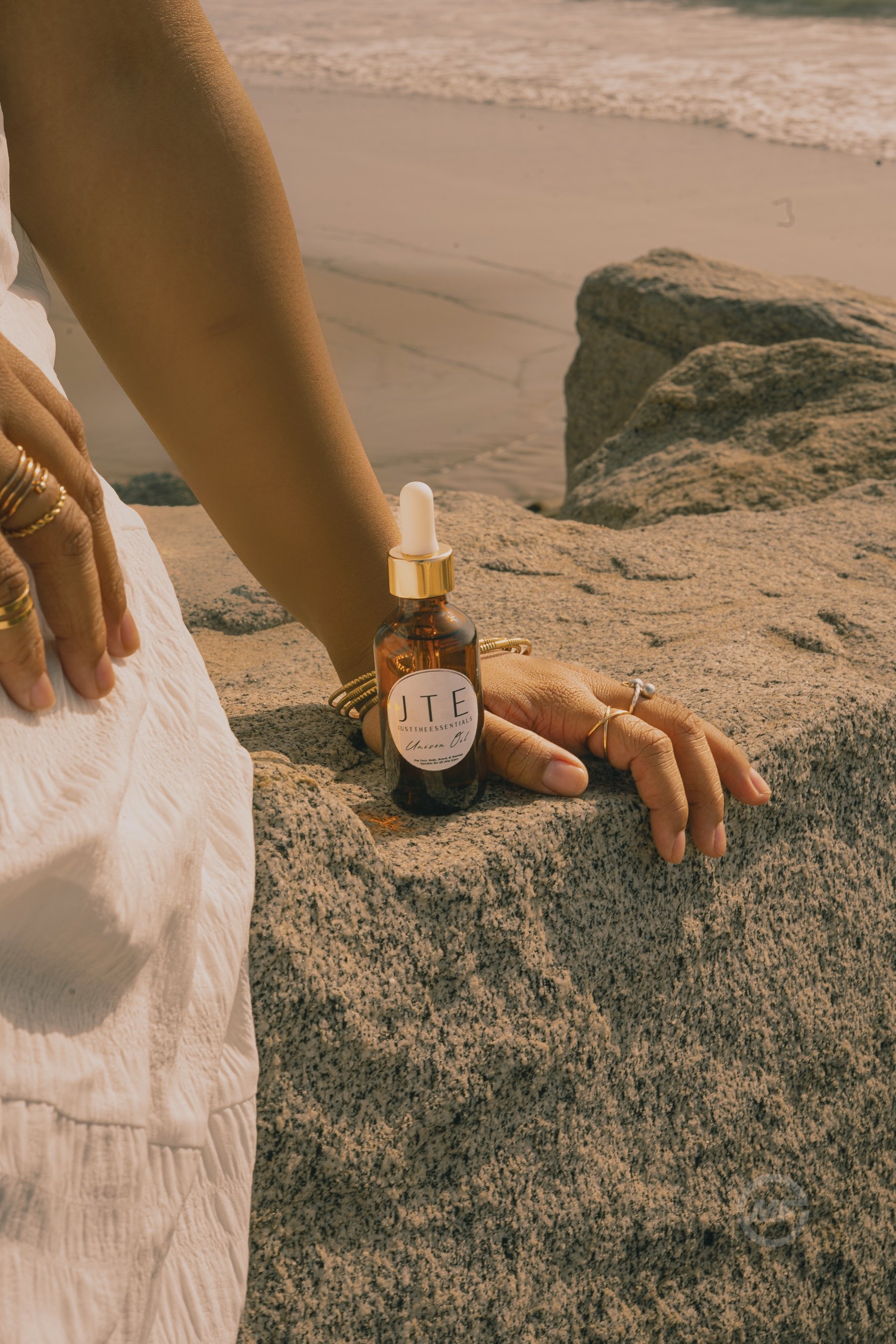 A person sitting on a rock by the beach with a small bottle of essential oil labeled 'JTE Justtheessentials Body Oil' placed on the rock near their hand, which is resting on the rock.