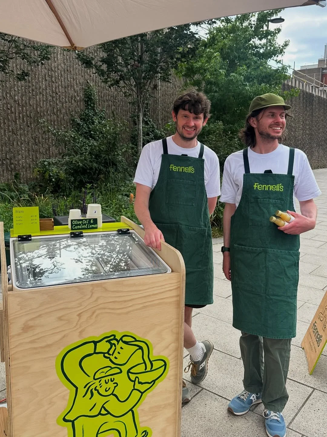 Two smiling men wearing green aprons with 'Fennell's' logo, standing next to a wooden ice cream cart outdoors, under a parasol, with green trees in the background.