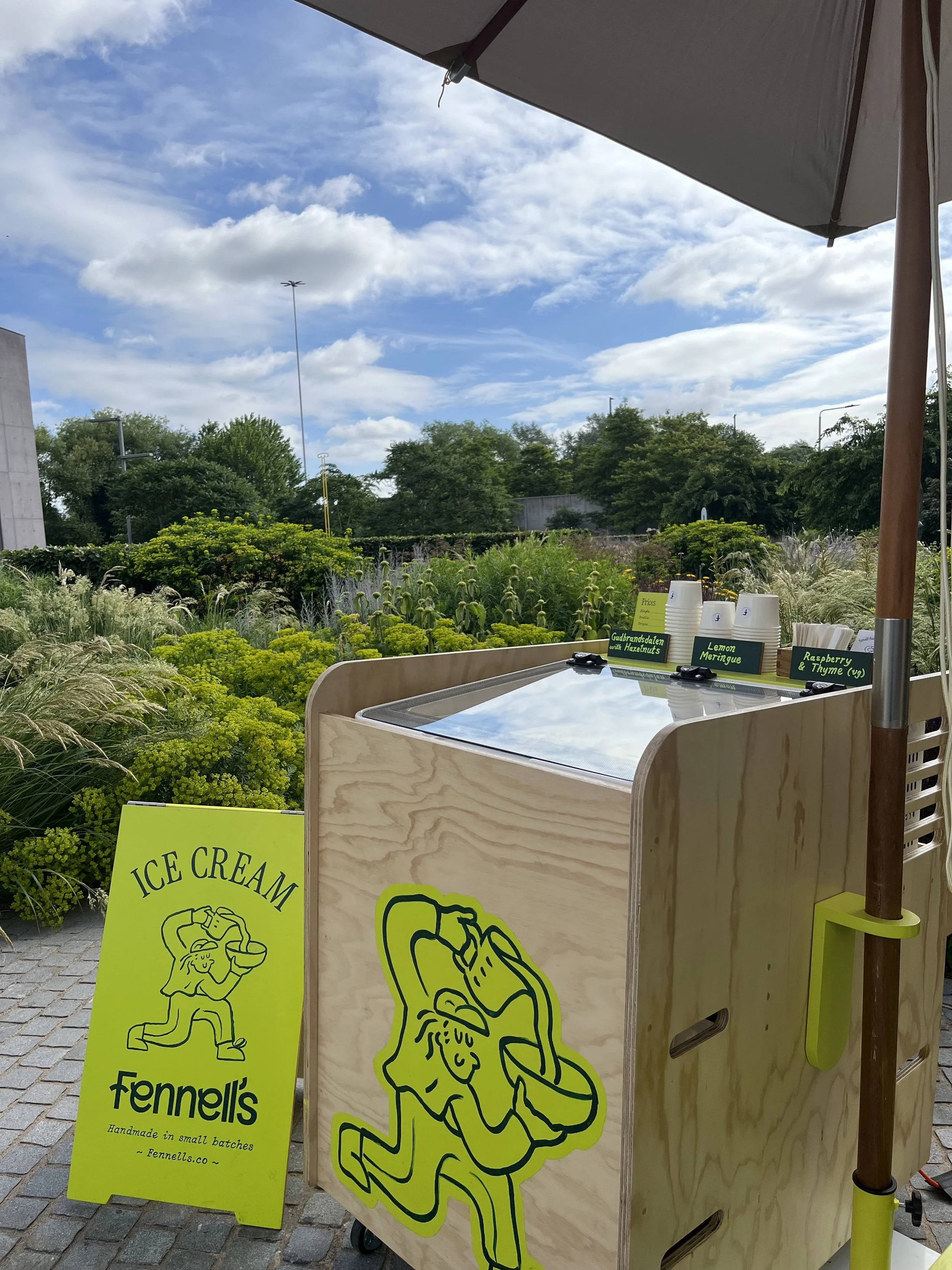 A mobile ice cream stand made featuring a neon yellow cartoon figure. The stand is set outdoors with lush green plants in the background.