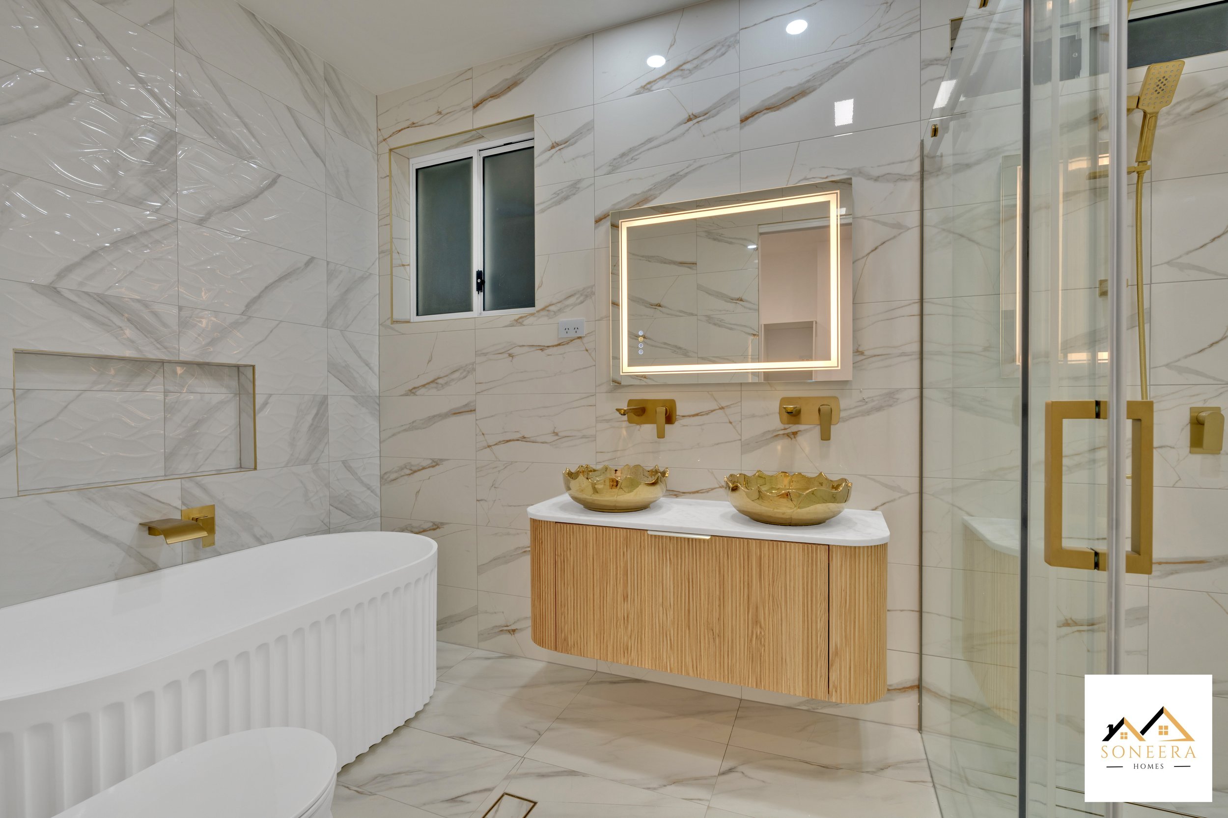 Modern bathroom with marble walls and floor, a white bathtub, a wooden vanity with two gold bowls, a backlit mirror, a small window, and a glass shower enclosure with gold handles.