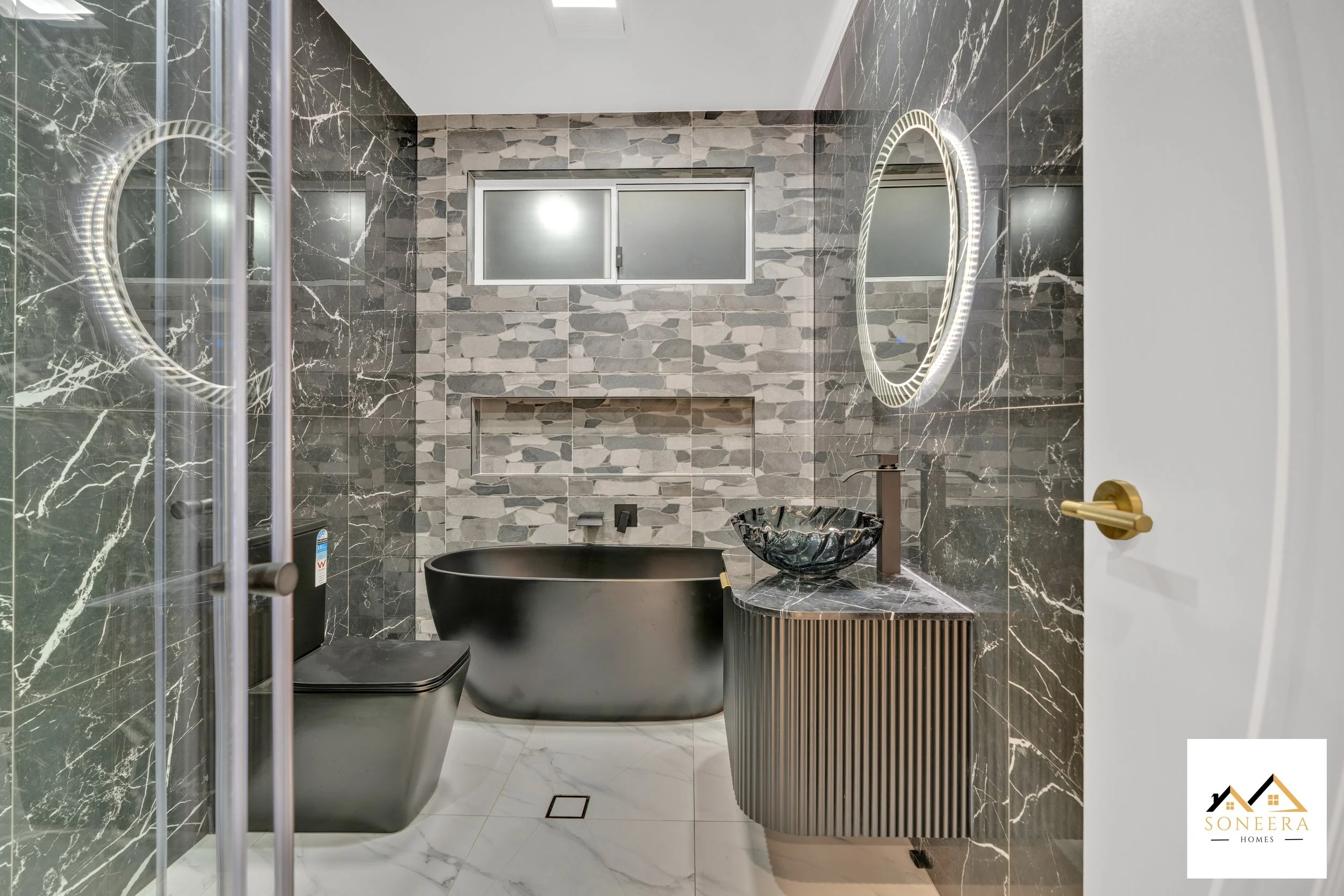 Modern bathroom with black countertop bathtub, black toilet, black marble sink, and circular LED mirror. Features gray stone wall tiles, a frosted window, and white marble floor.