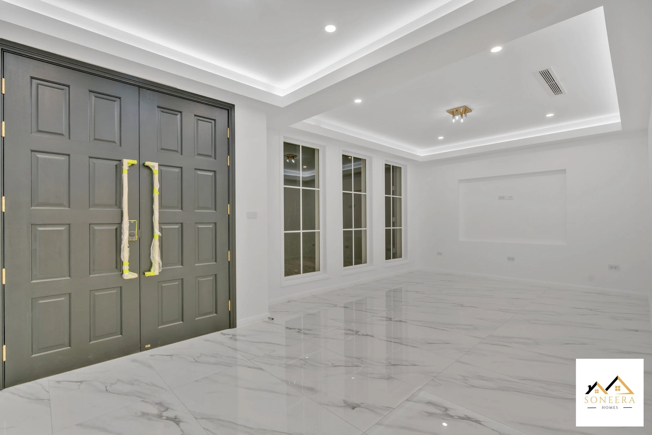 Empty modern living room with white walls, marble tile flooring, large glass windows, and a grey double-door entrance. Recessed lighting and a ceiling with cove lighting detail.
