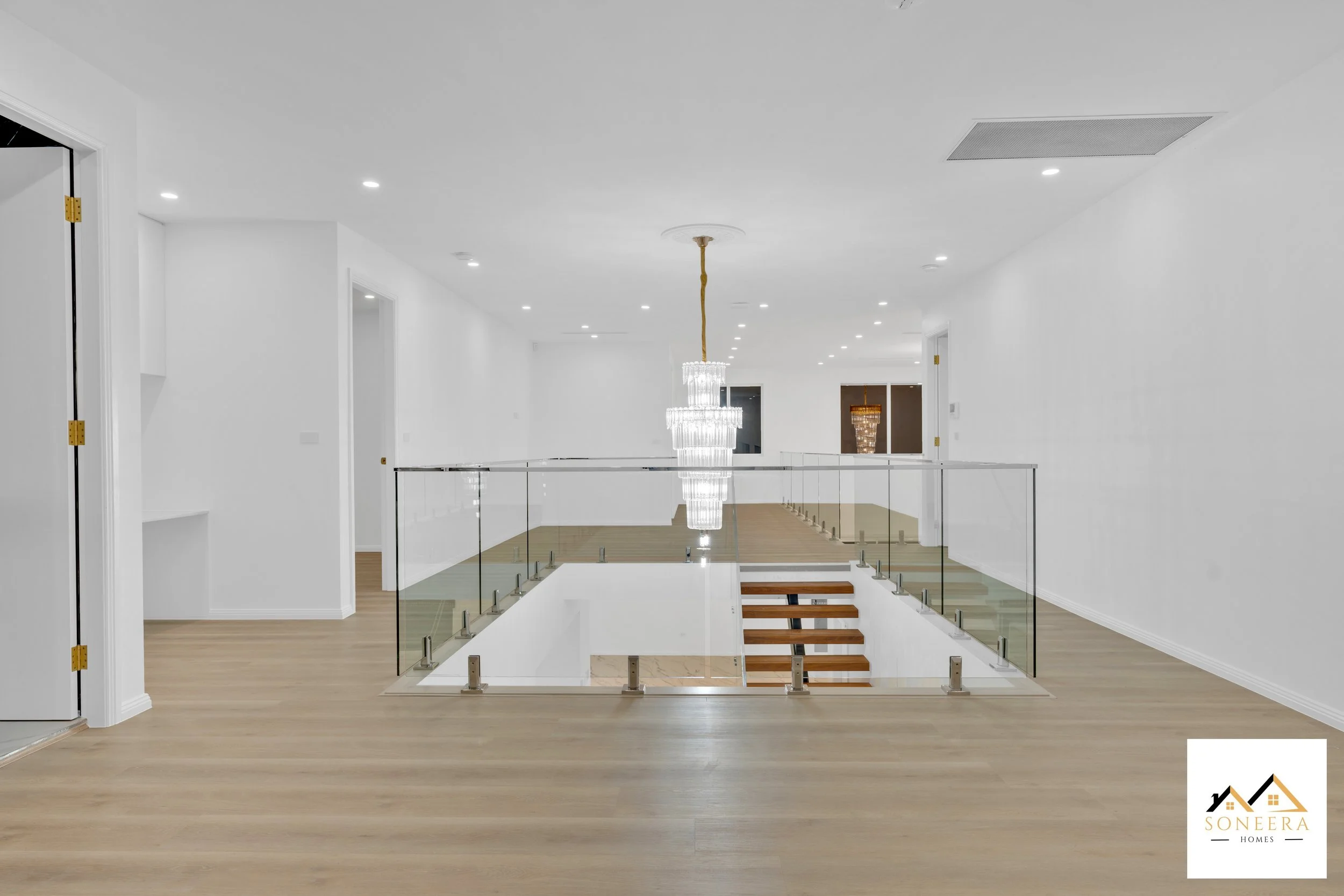 Interior of a modern, minimalist home featuring a staircase with glass railings and a large chandelier hanging from the ceiling. The room has white walls, recessed lighting, and light wooden flooring.