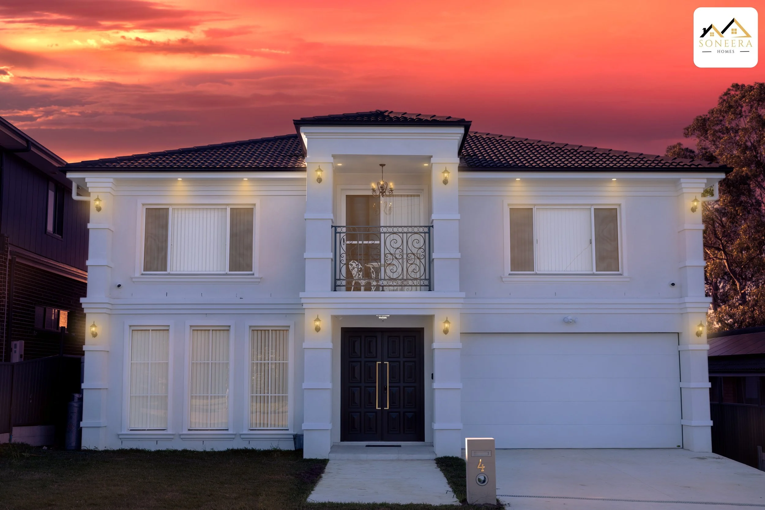 A modern white two-story house at sunset with a black front door, multiple windows with blinds, small balcony with wrought iron railing, exterior lights, and a driveway.