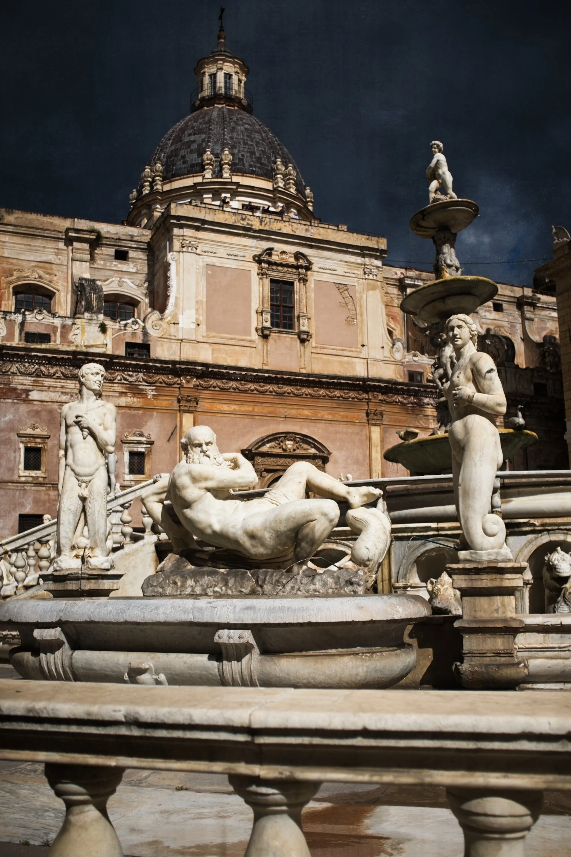 A historic European fountain with marble statues of mythological figures in front of an old building with a large dome.