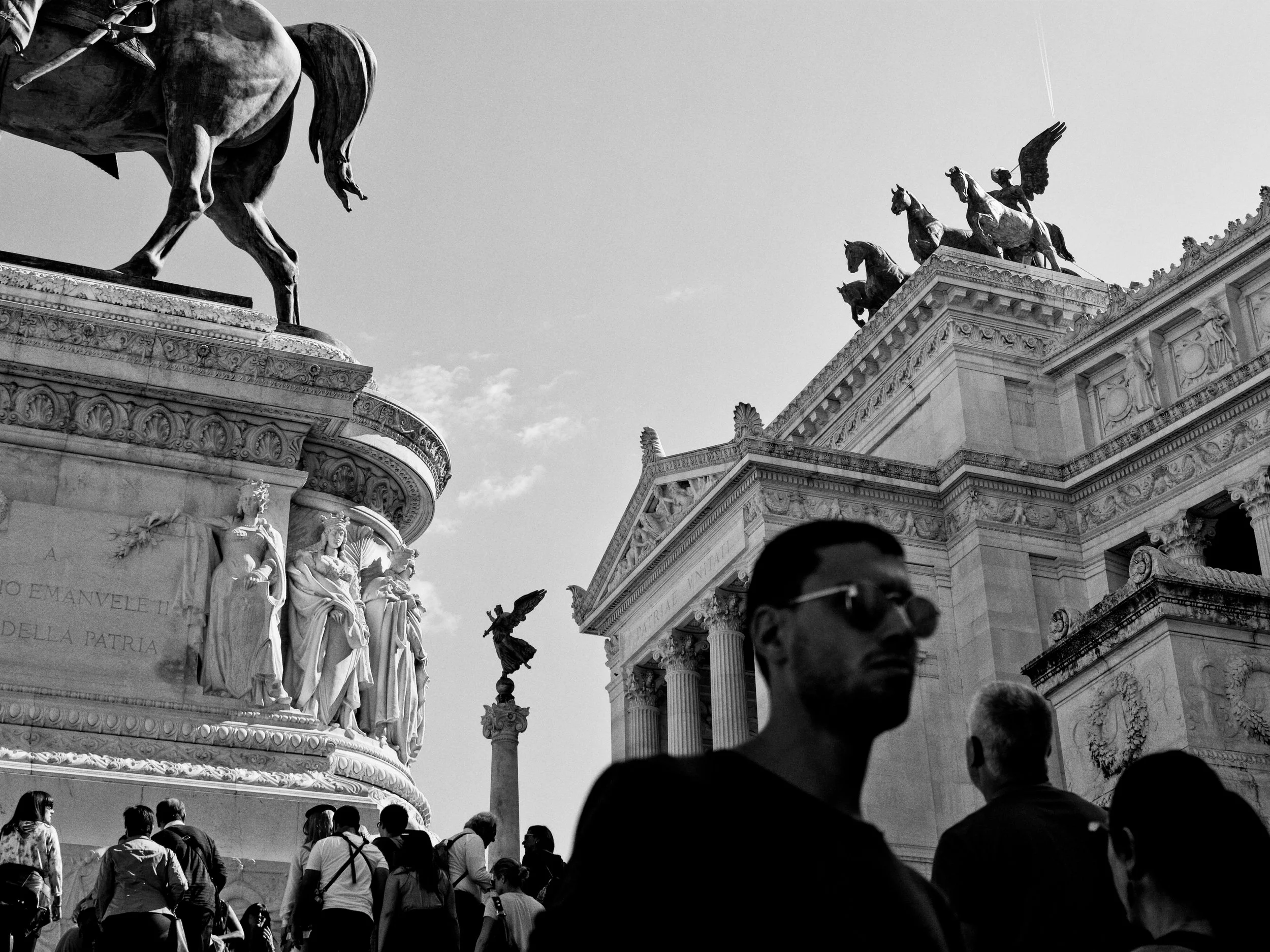 People walking in front of classical European architecture with statues and sculptures, including a chariot with horses and winged figures, in a city square.