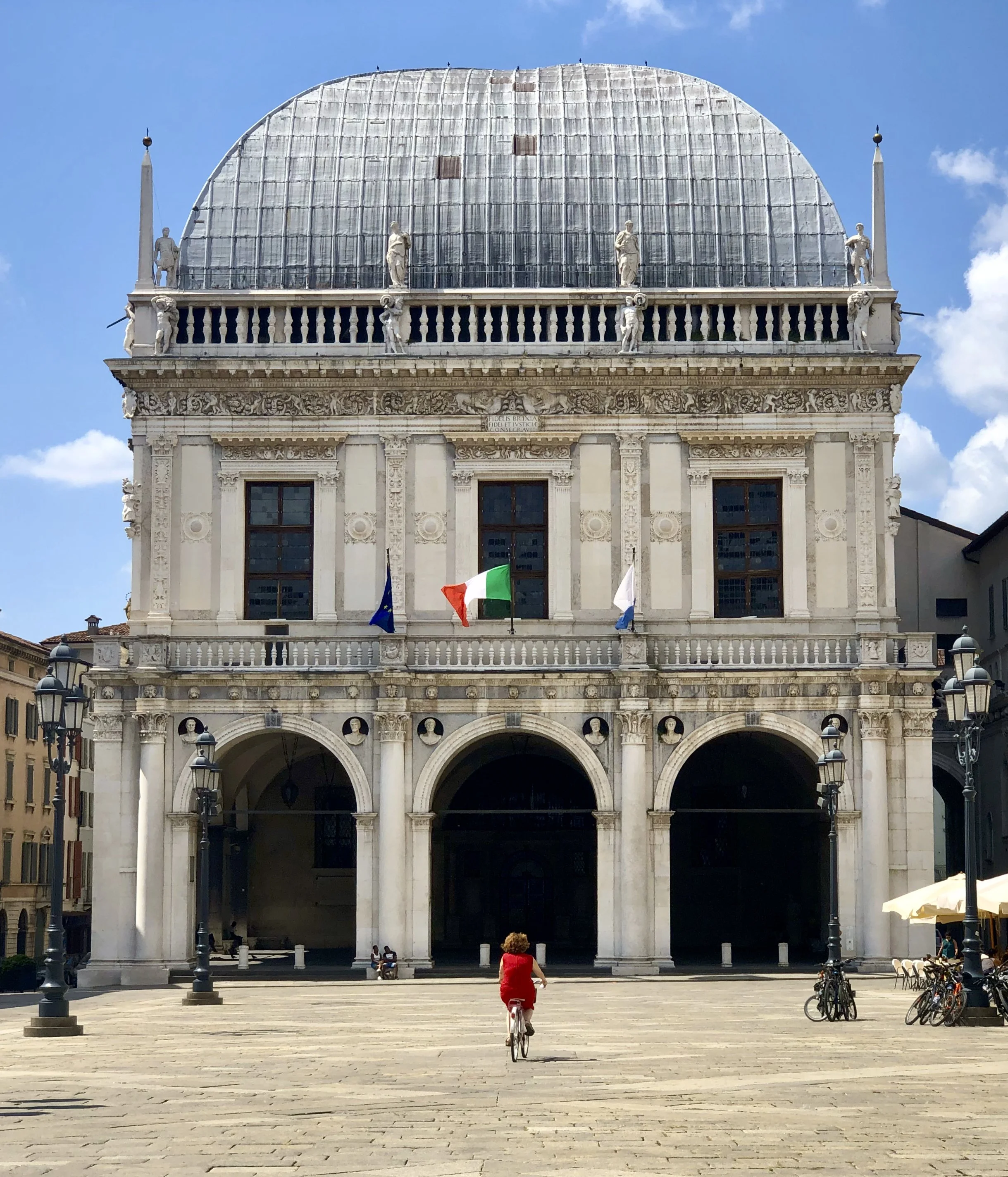 A historic building with classical architecture, featuring three archways at the entrance, ornate decorations, and a curved glass dome on top. Flags of France, the European Union, and a white flag are displayed in front. In the foreground, a child in