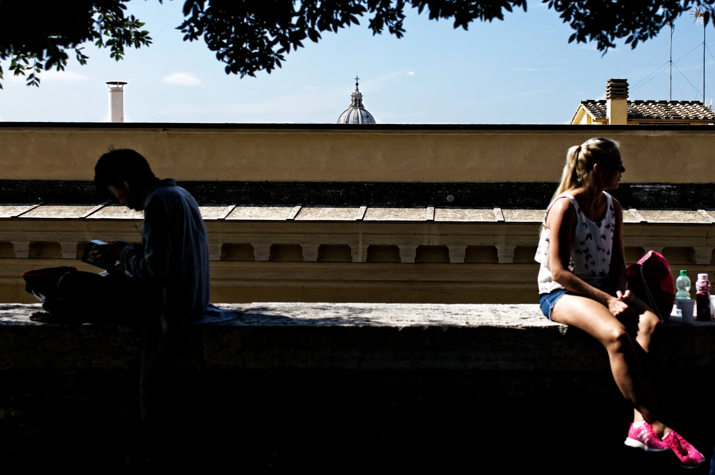 A silhouette of a man sitting on a stone ledge, using a phone, and a woman sitting on the same ledge with her back to the man, wearing sunglasses and pink sneakers, with water bottles and a backpack beside her, under a tree with buildings and a domed