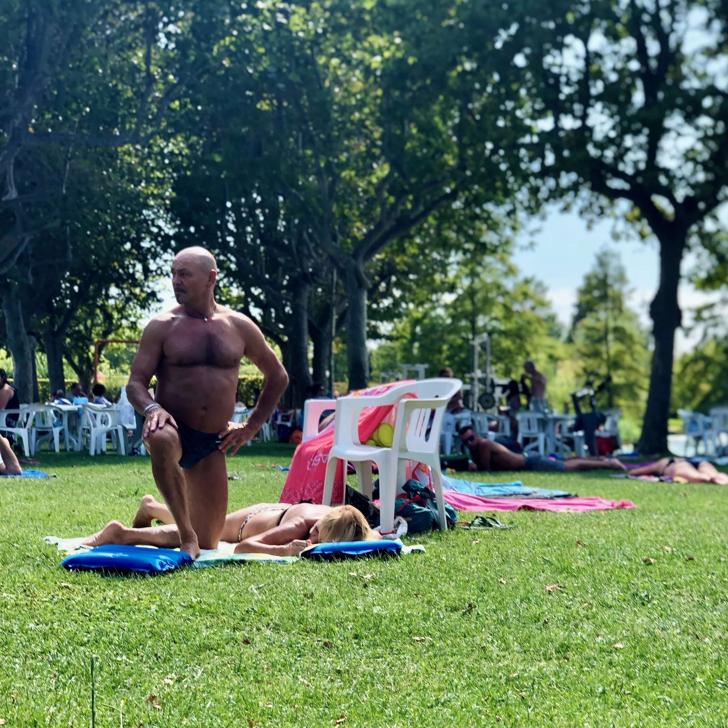 A shirtless man in black shorts is kneeling on a towel on grass, while a woman with blonde hair lies face down on a blanket nearby in a park with trees. Several other people are visible in the background relaxing and sitting among white chairs and um
