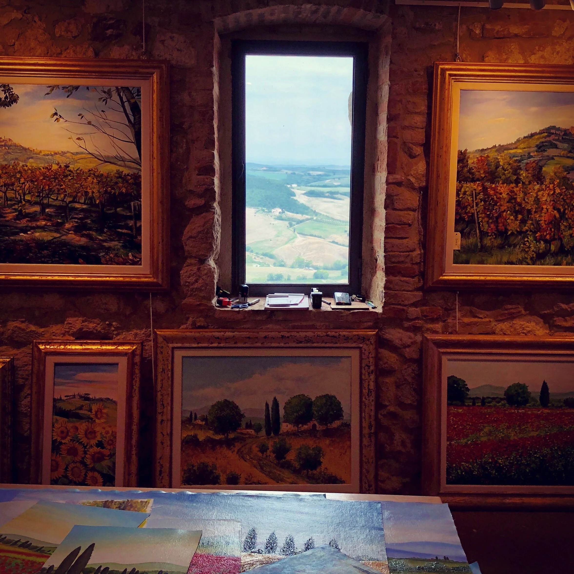 An art gallery with landscape paintings on a stone wall, a window showing a scenic view of rolling hills and fields, and art supplies on a table in the foreground.