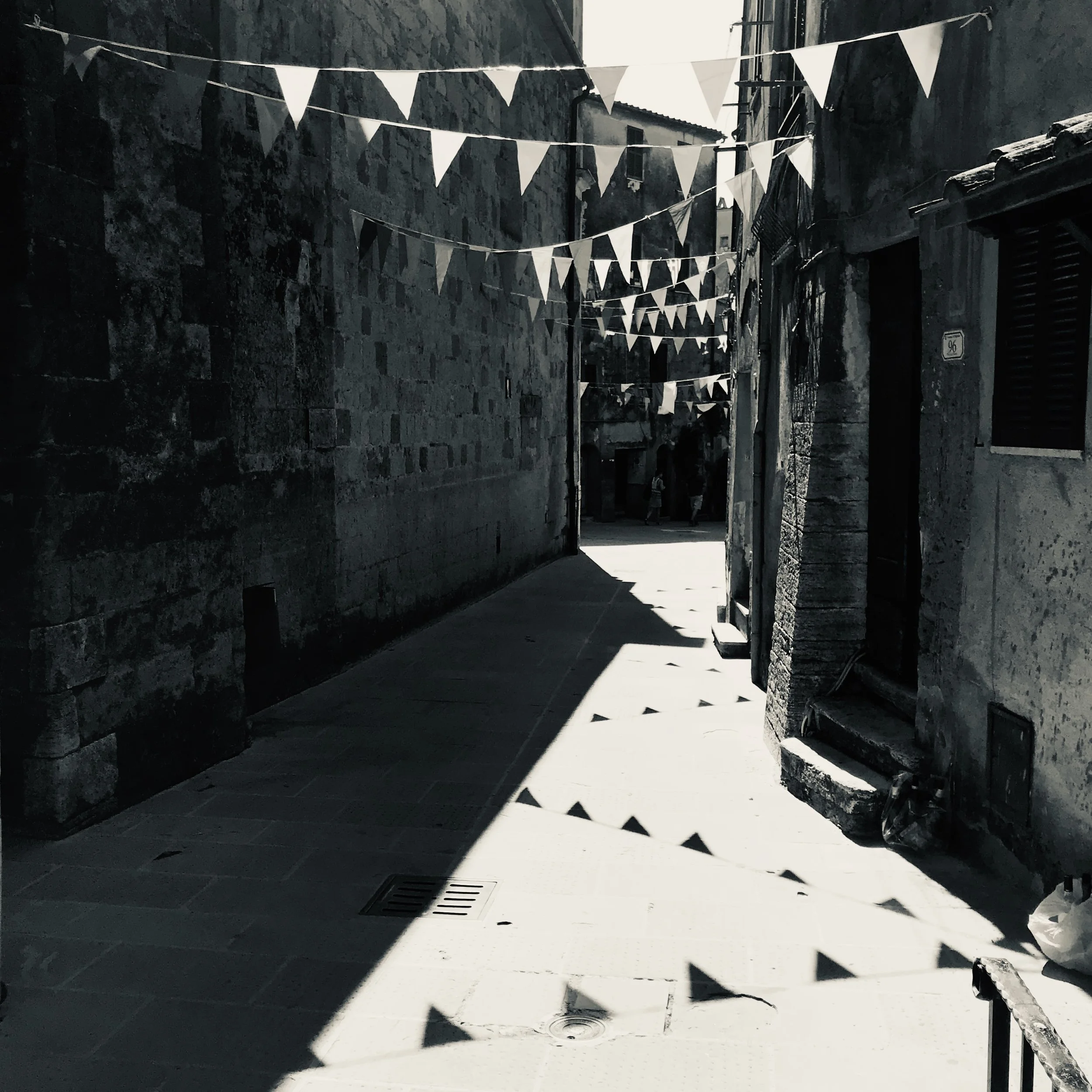 Narrow stone-paved alley with hanging bunting flags and shadows cast from buildings, in sunlight.