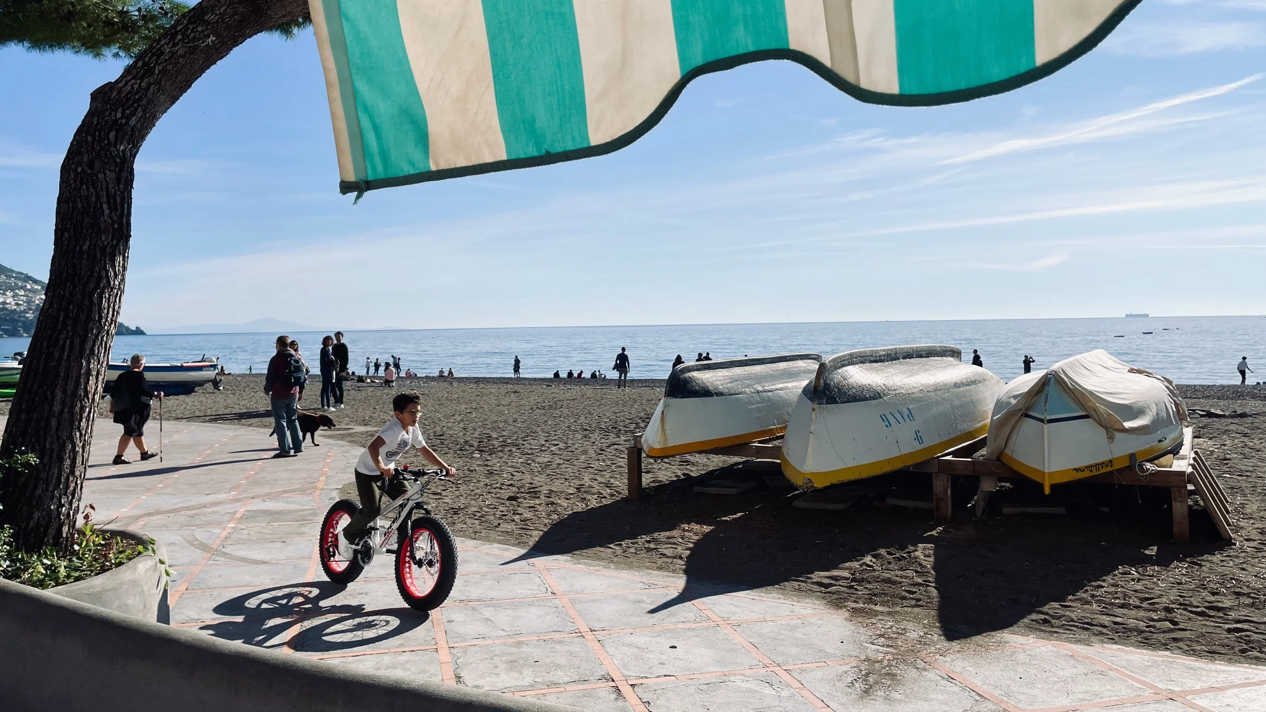 Children and adults on a beach promenade with boats and the ocean in the background. A boy rides a bicycle while others walk or stand near the shoreline. Several boats are turned over on the sand. A striped beach umbrella is partially visible at the 