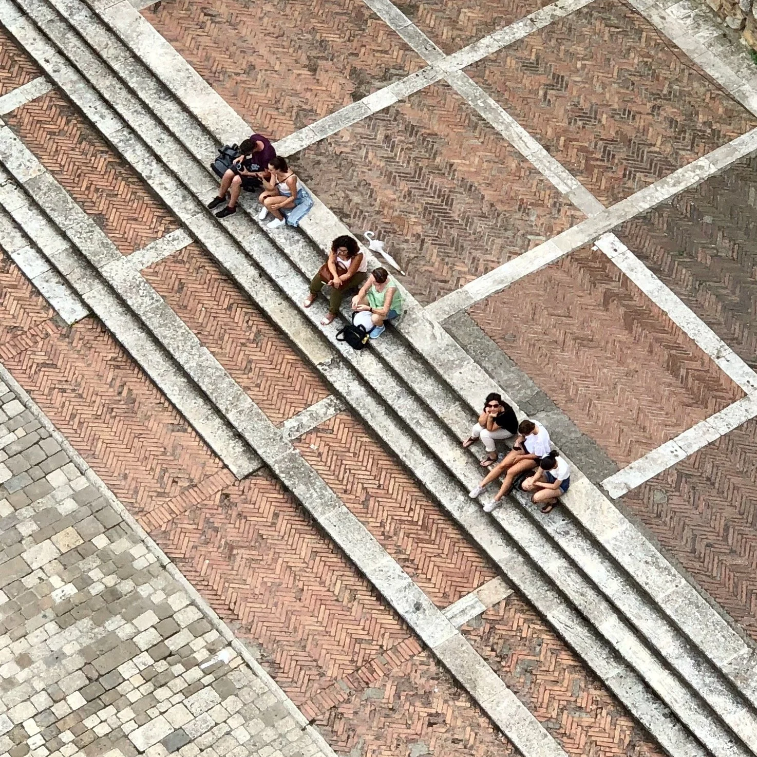 Bird's-eye view of seven people sitting on stone steps on a brick pedestrian area, with some holding umbrellas and bags.