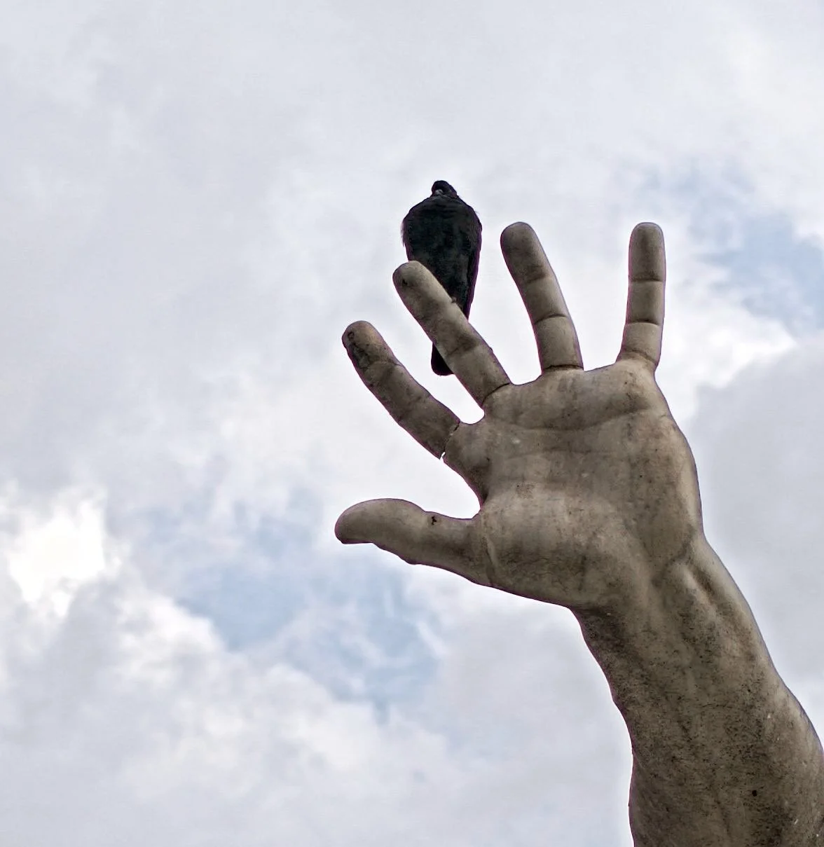 A raised hand sculpture reaching towards a gray, cloudy sky, with a black bird perched on the index finger.