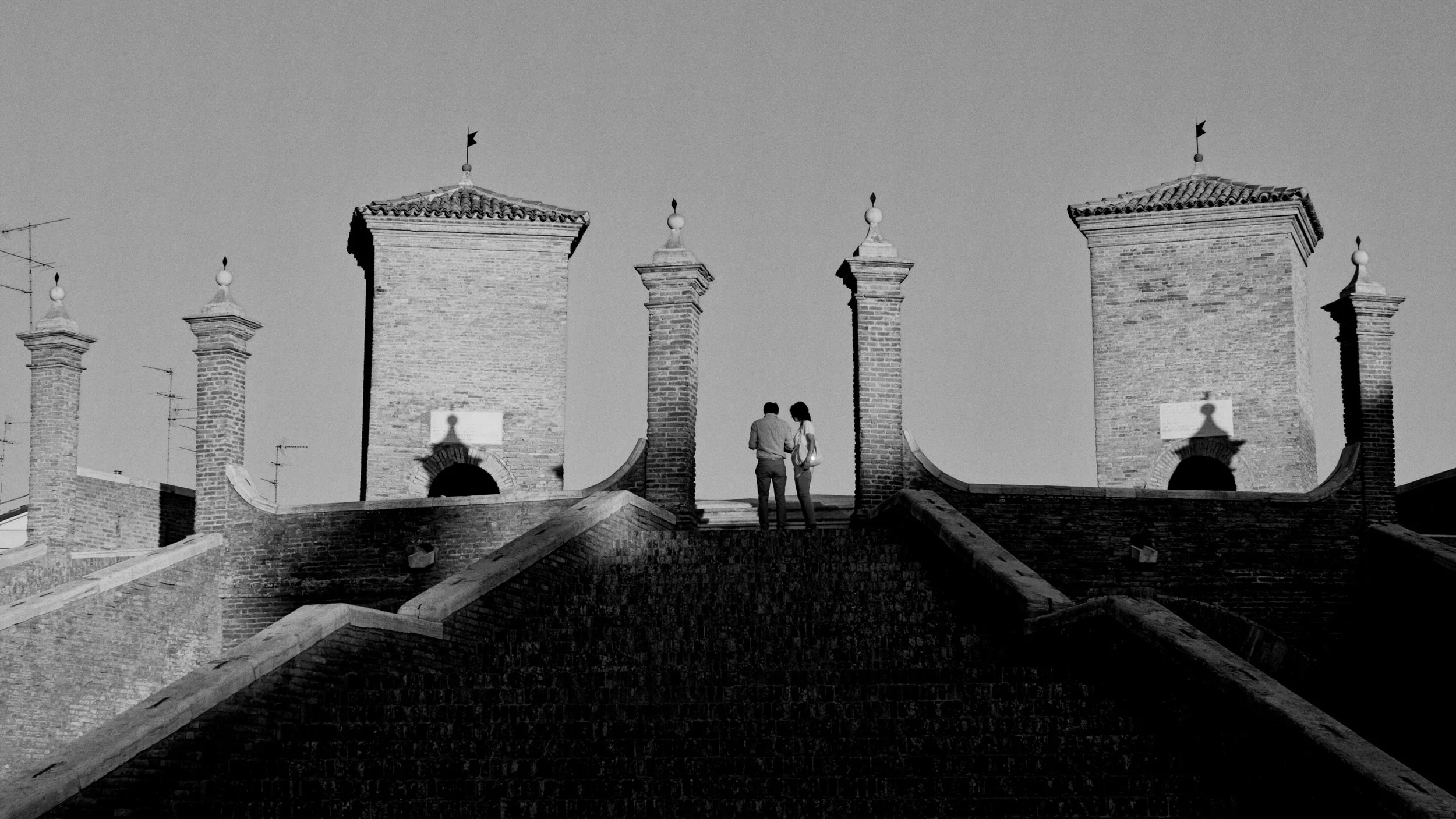 Black and white photo of two people standing on a rooftop, surrounded by brick chimneys with tiled roofs, under a clear sky.