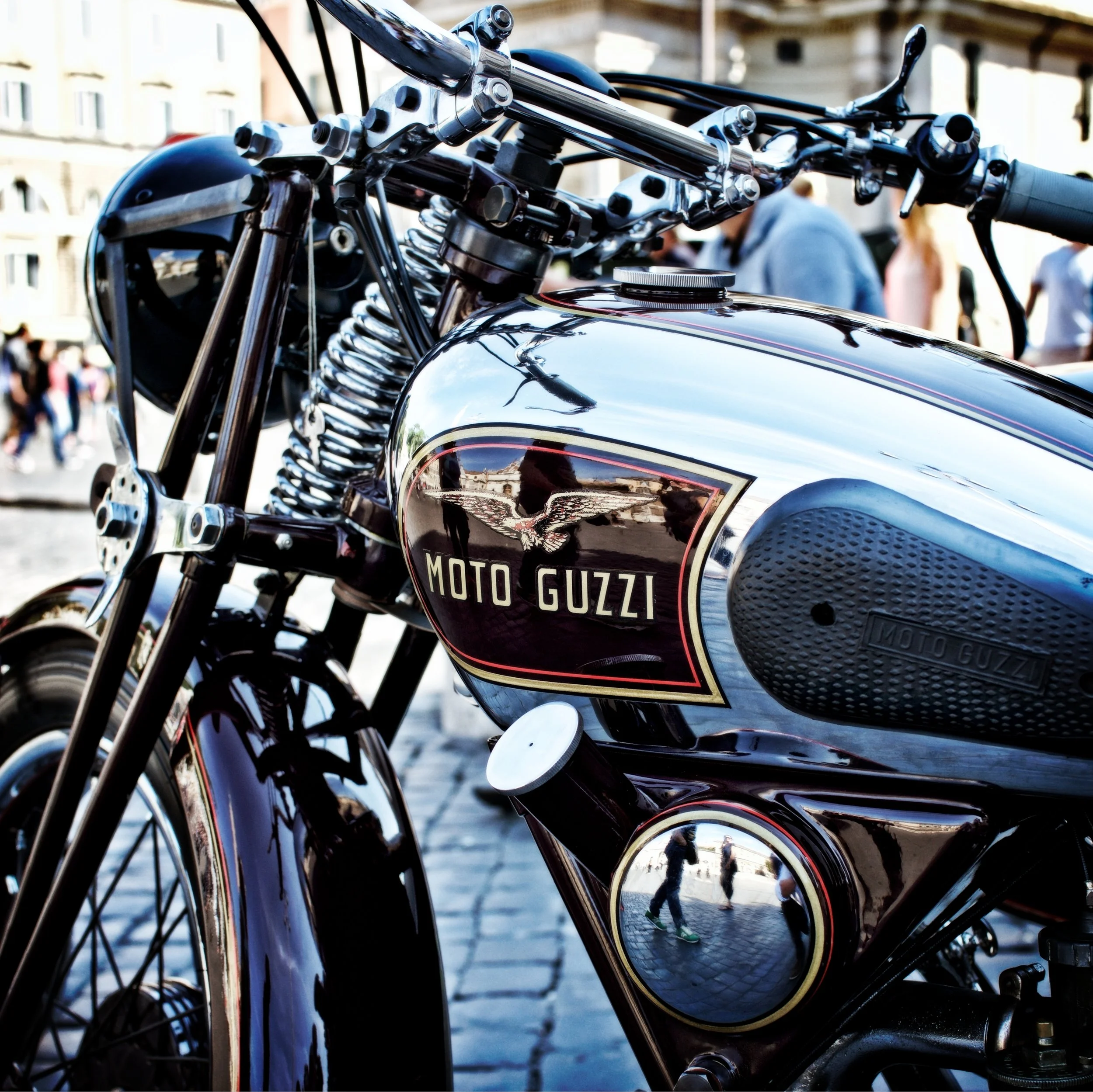 Close-up of a Moto Guzzi motorcycle with chrome details and a black leather seat, parked outdoors with people and buildings in the background.