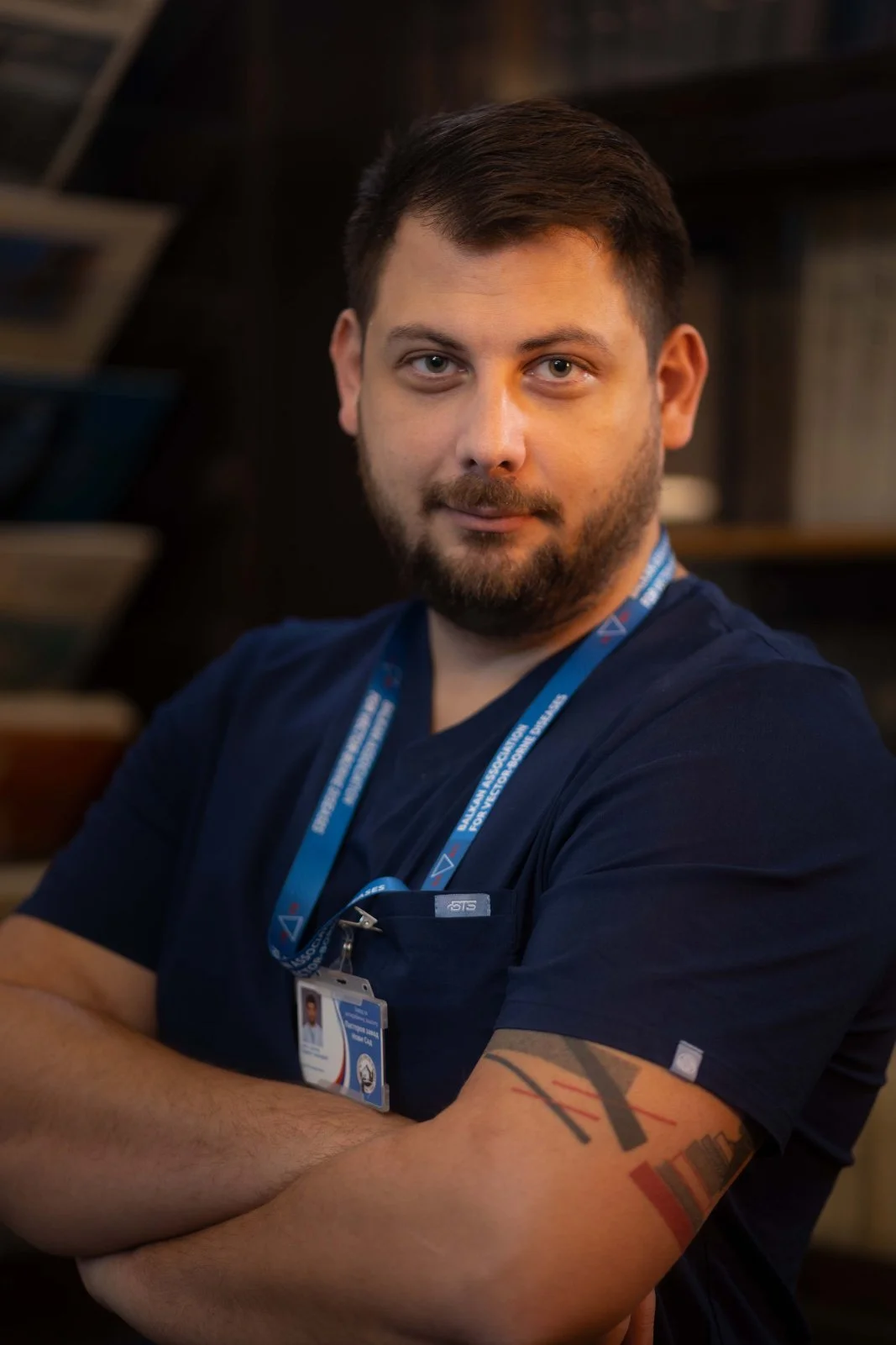 A male healthcare professional wearing blue scrubs and a name badge, standing with arms crossed in a clinical setting.
