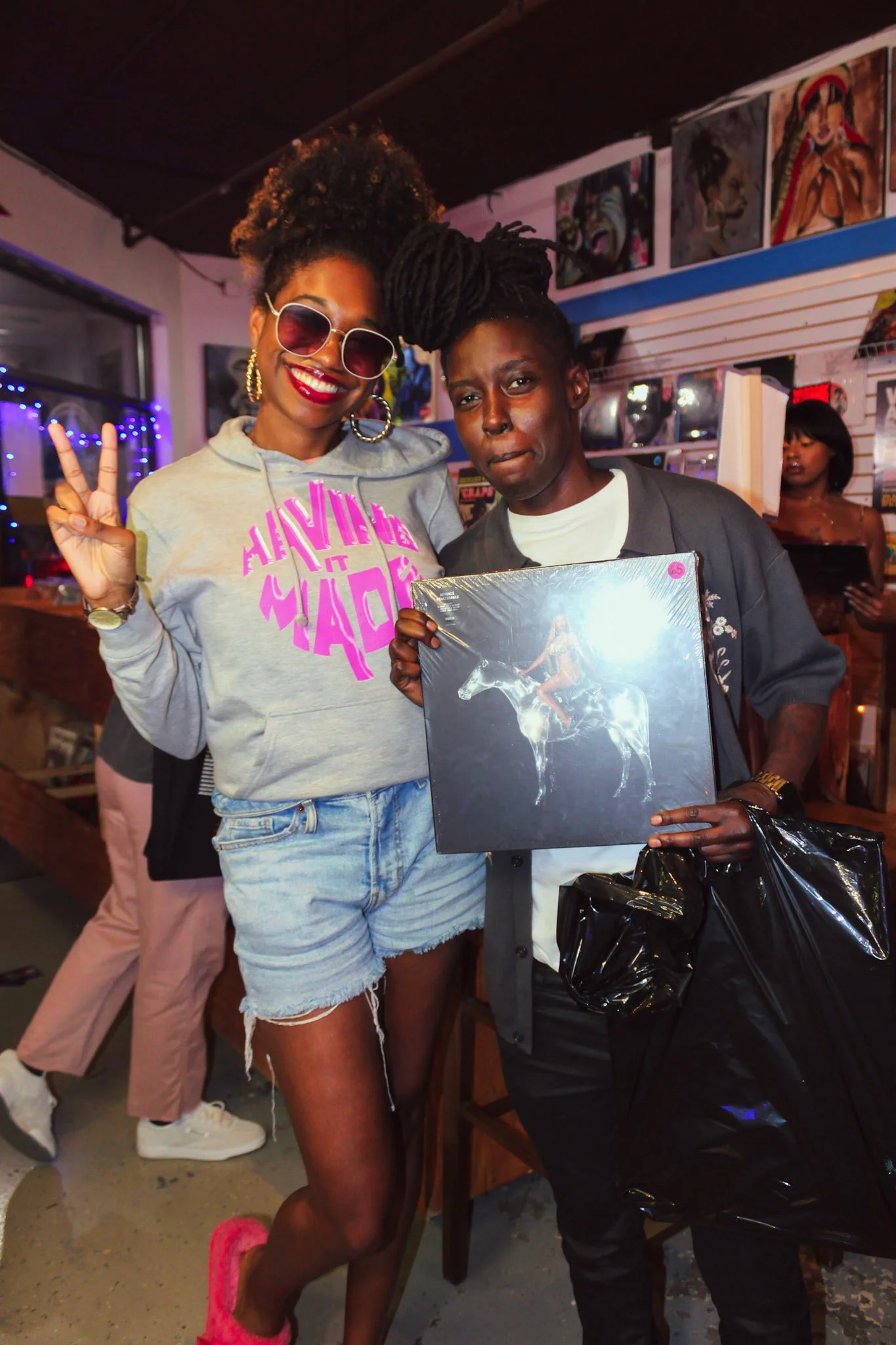 Two women posing inside a record store, one making a peace sign and the other holding a vinyl record with a depiction of a woman riding a horse. The woman on the left is wearing sunglasses, a sweatshirt, and denim shorts, while the woman on the right is holding a black shopping bag and wearing a dark jacket.