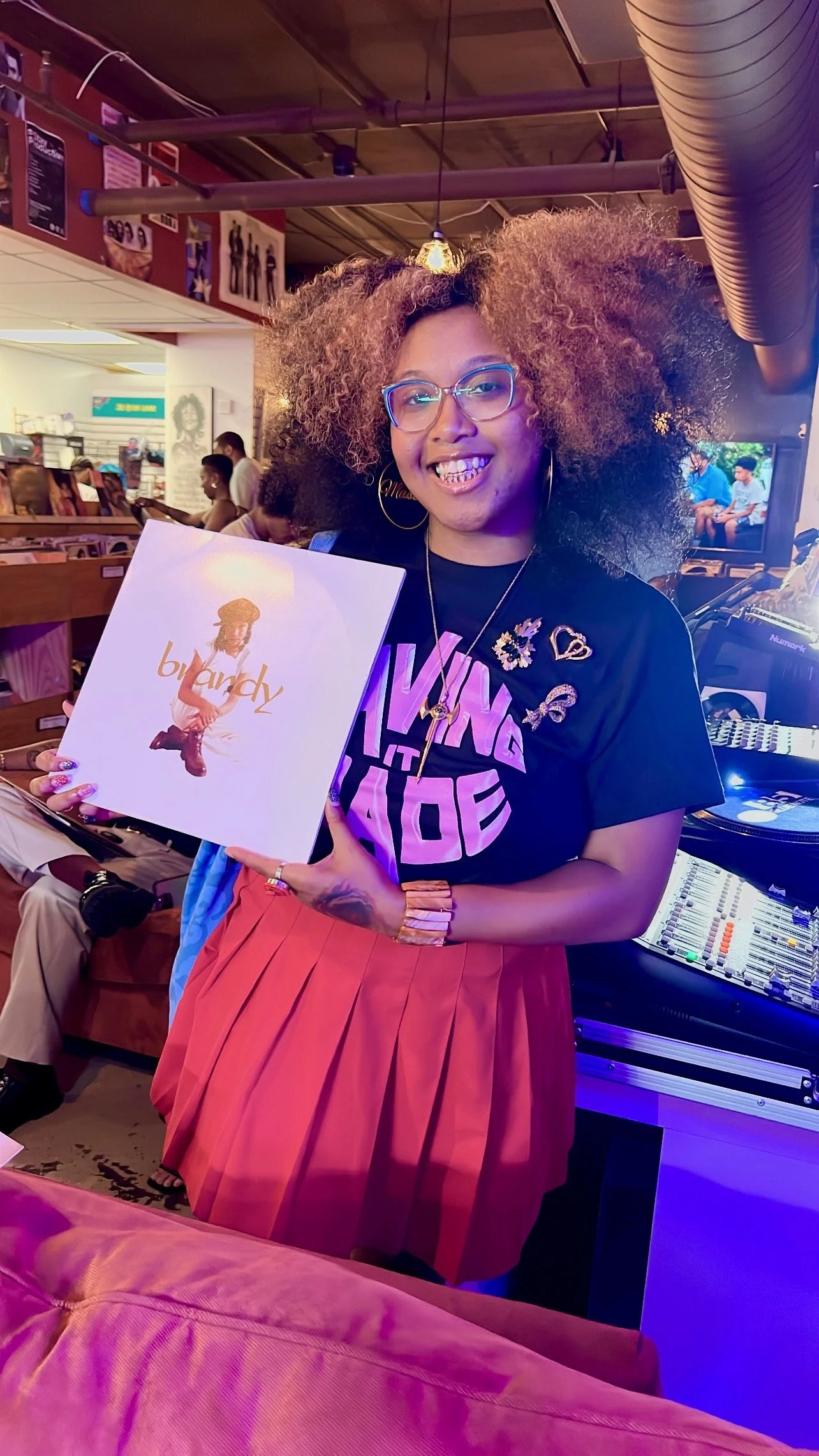 A woman with curly hair, glasses, and a big smile holding a vinyl record with her photo on the cover. She is wearing a black T-shirt with pink text and a red pleated skirt, standing in a record store or music venue.