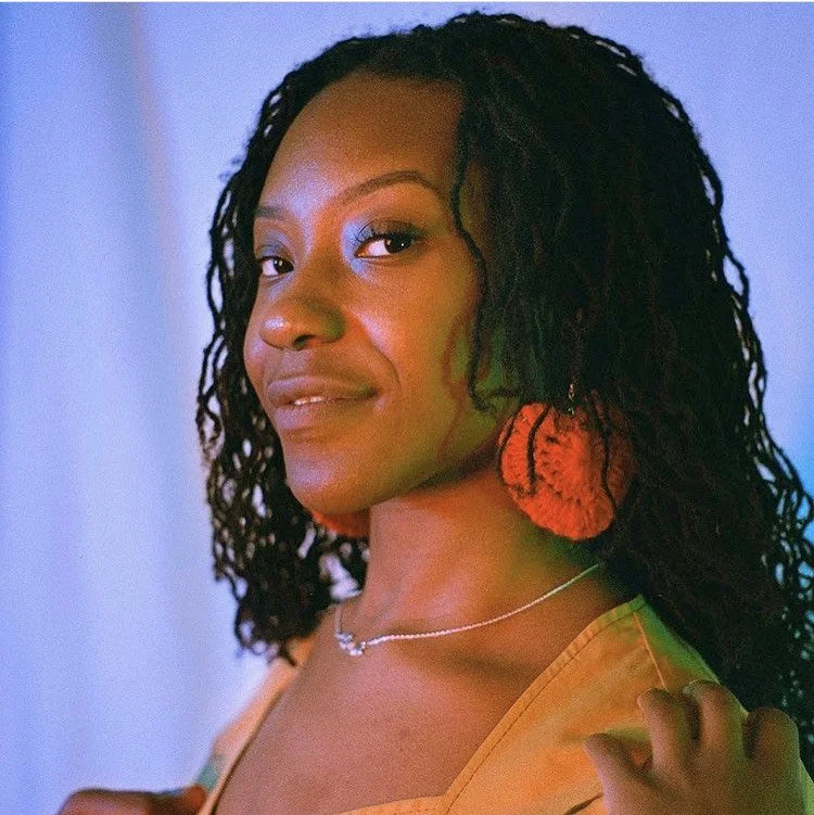 A woman with curly black hair wearing orange earrings and a necklace, smiling gently, with a light-colored top against a soft blue and purple background.