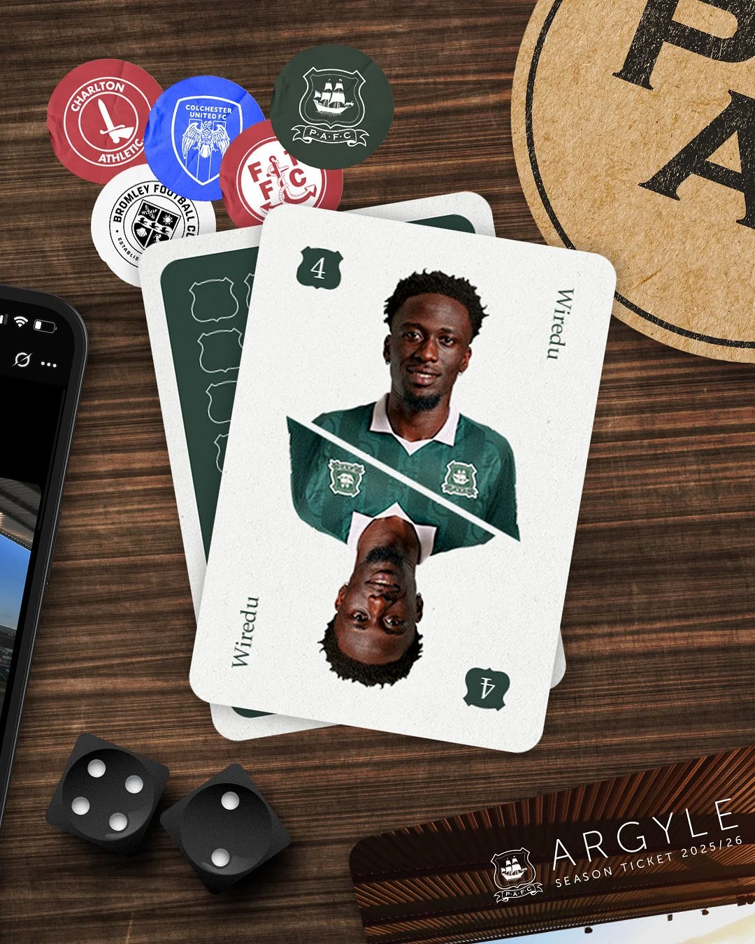 A deck of soccer cards featuring a player from Plymouth Argyle, with one card showing a portrait of the player labeled as 'Wired.' The cards are on a wooden surface alongside scattered black dice, soccer club badges, and a smartphone, with a coffee cup paper coaster partially visible.