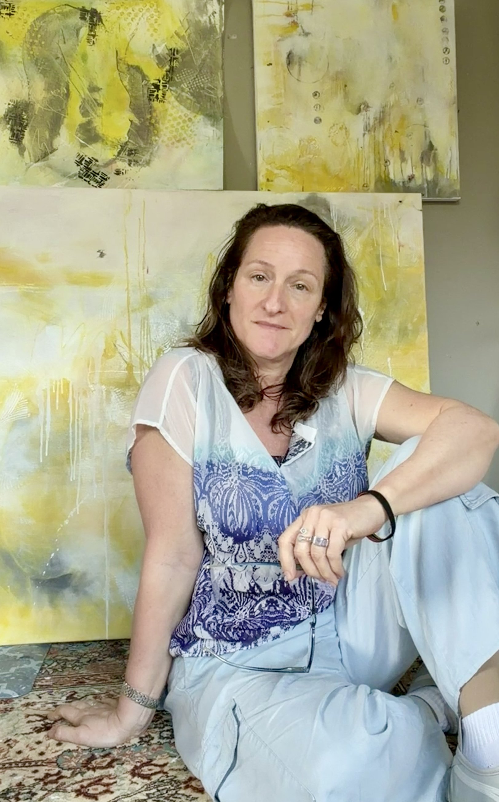 A woman with shoulder-length reddish-brown hair smiling in front of abstract artwork and a wall. She is wearing hoop earrings and a black top.