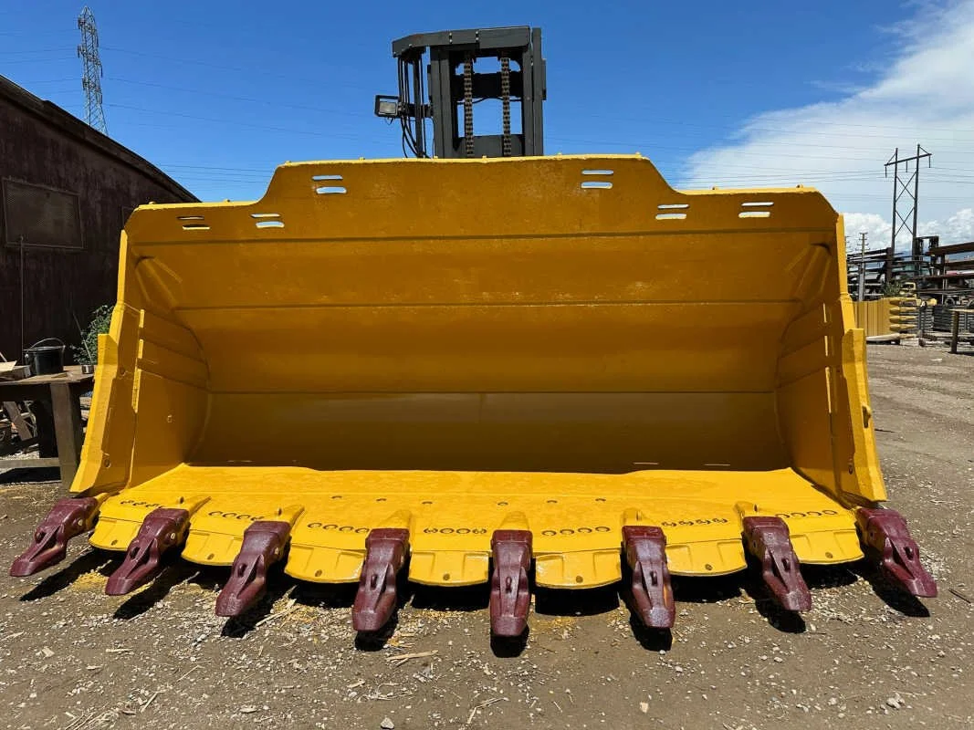 A large yellow excavator bucket attachment with red teeth, placed outdoors against a blue sky with some clouds and industrial structures in the background.
