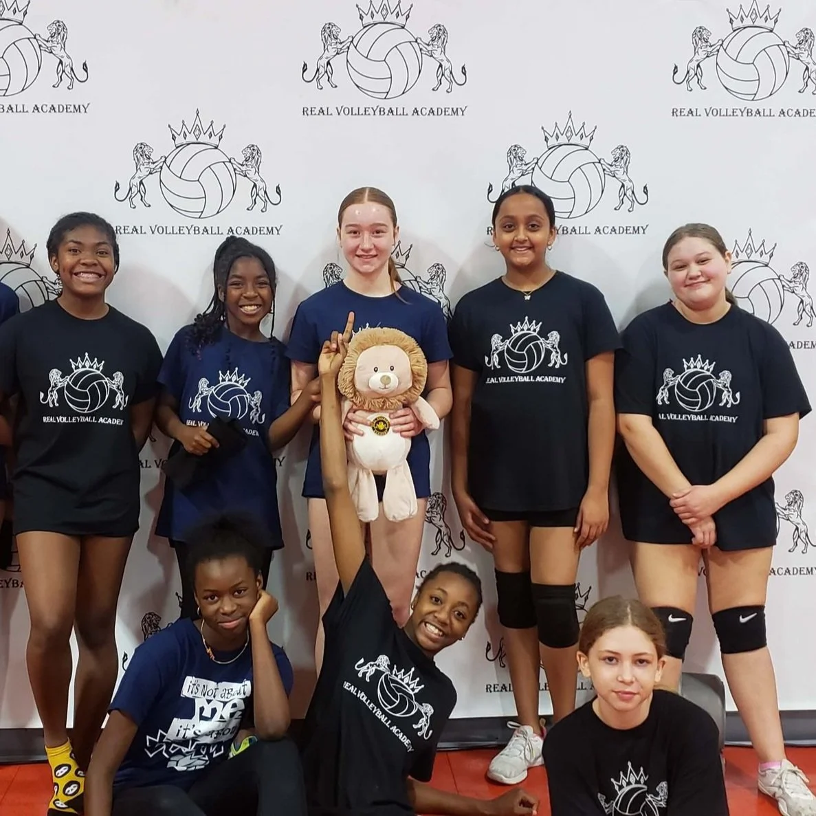 A group of young volleyball players wearing matching t-shirts from a volleyball academy stand and kneel in front of a backdrop with the academy's logo. One player is holding a stuffed toy lion.