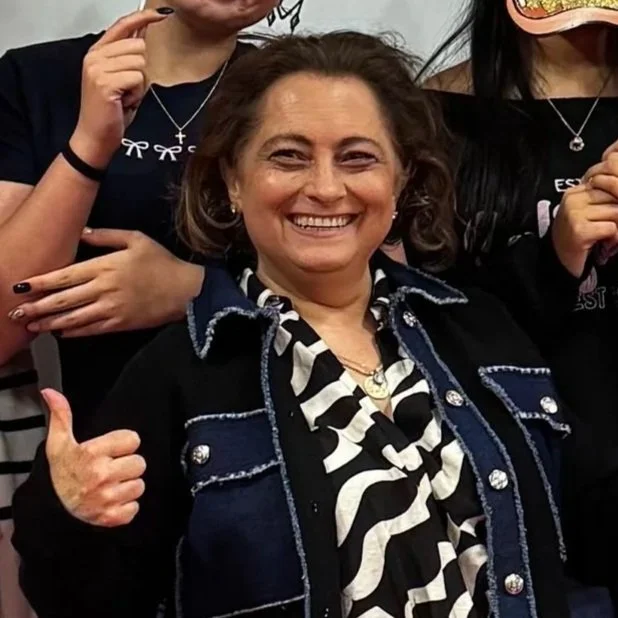 A woman with curly brown hair wearing a blue statement necklace and pink top, smiling slightly.