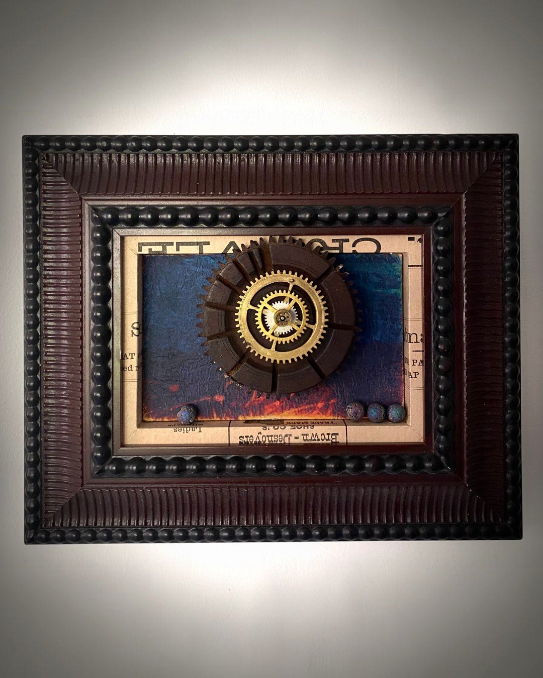 Assemblage artwork featuring concentric vintage gears against layered blue and black textured surfaces.