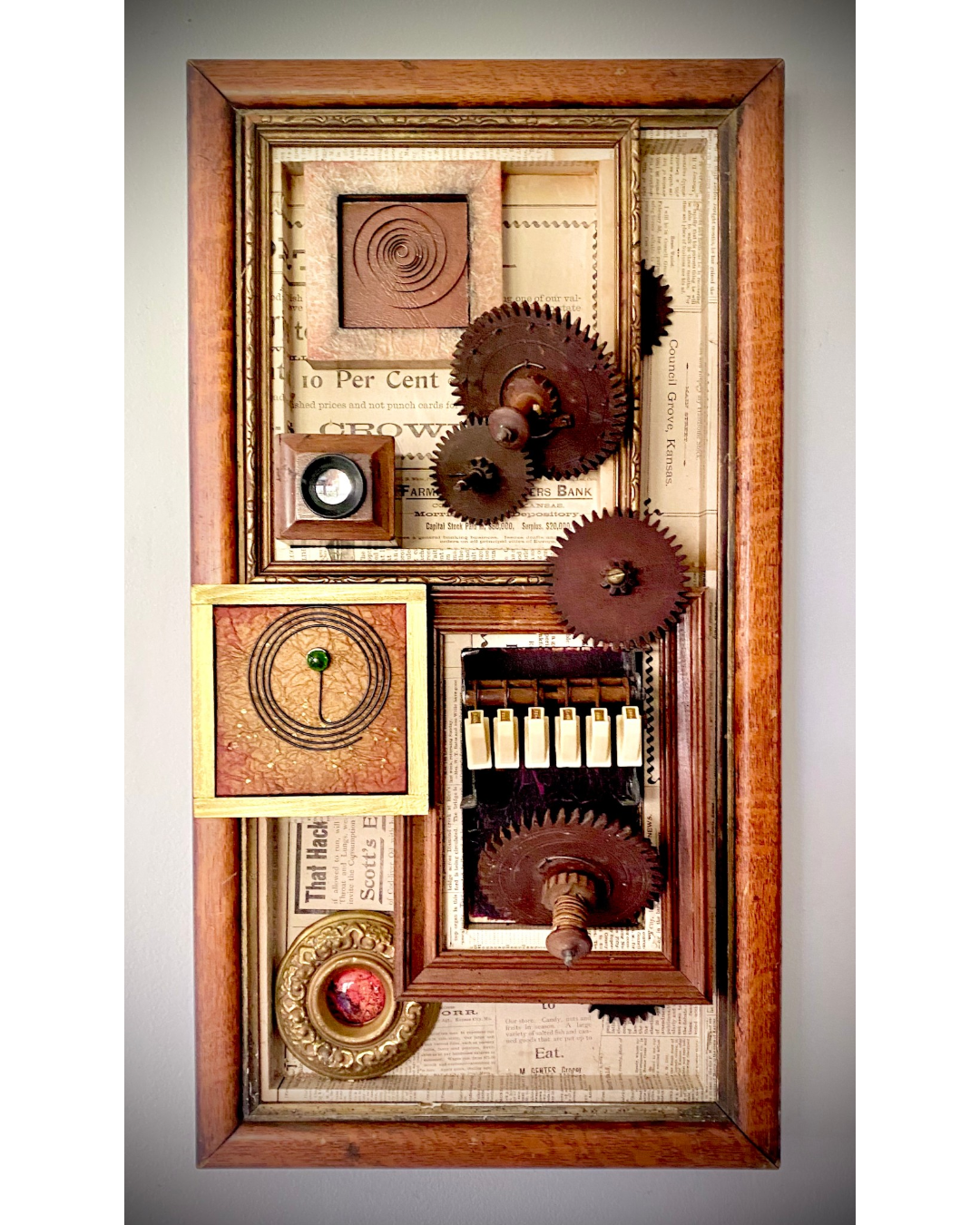 Assemblage artwork featuring interlocking vintage gears and layered printed papers within a shadow box frame.