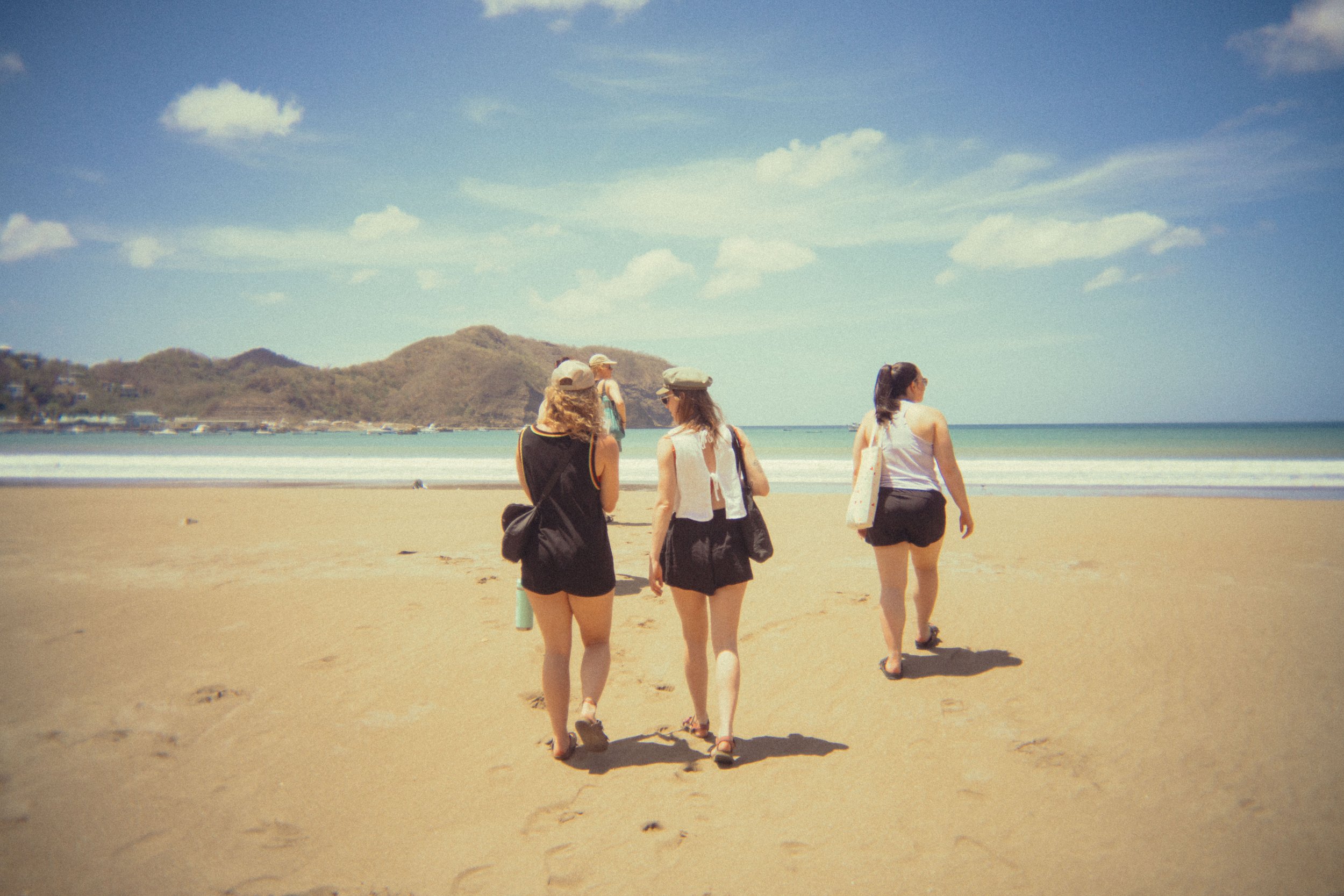 Four women walking on a sandy beach with mountains in the background and a blue sky with some clouds.