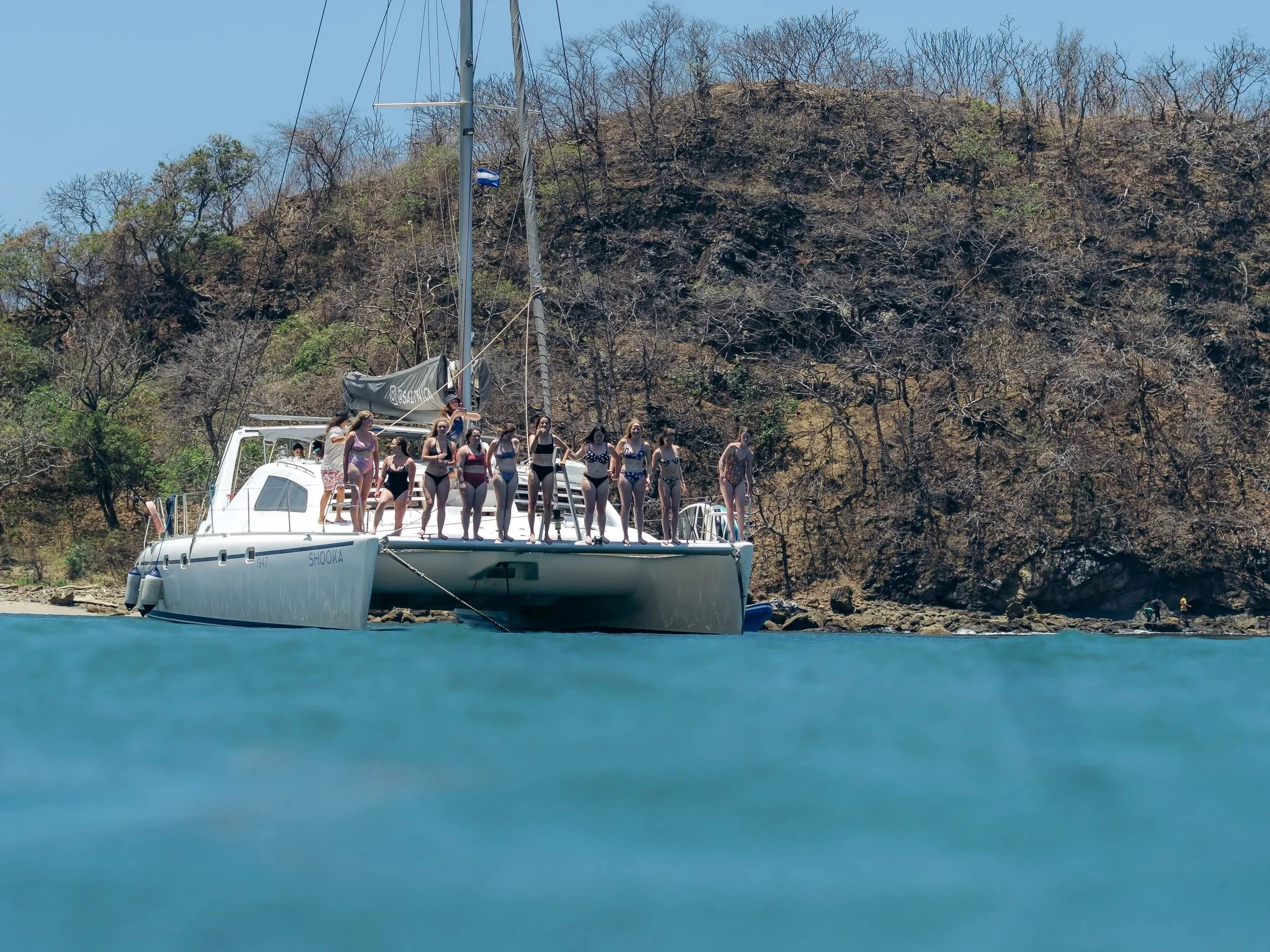 People standing on a sailboat named 'Shooka' anchored near a rocky shoreline with dry trees and a clear blue sky in the background.
