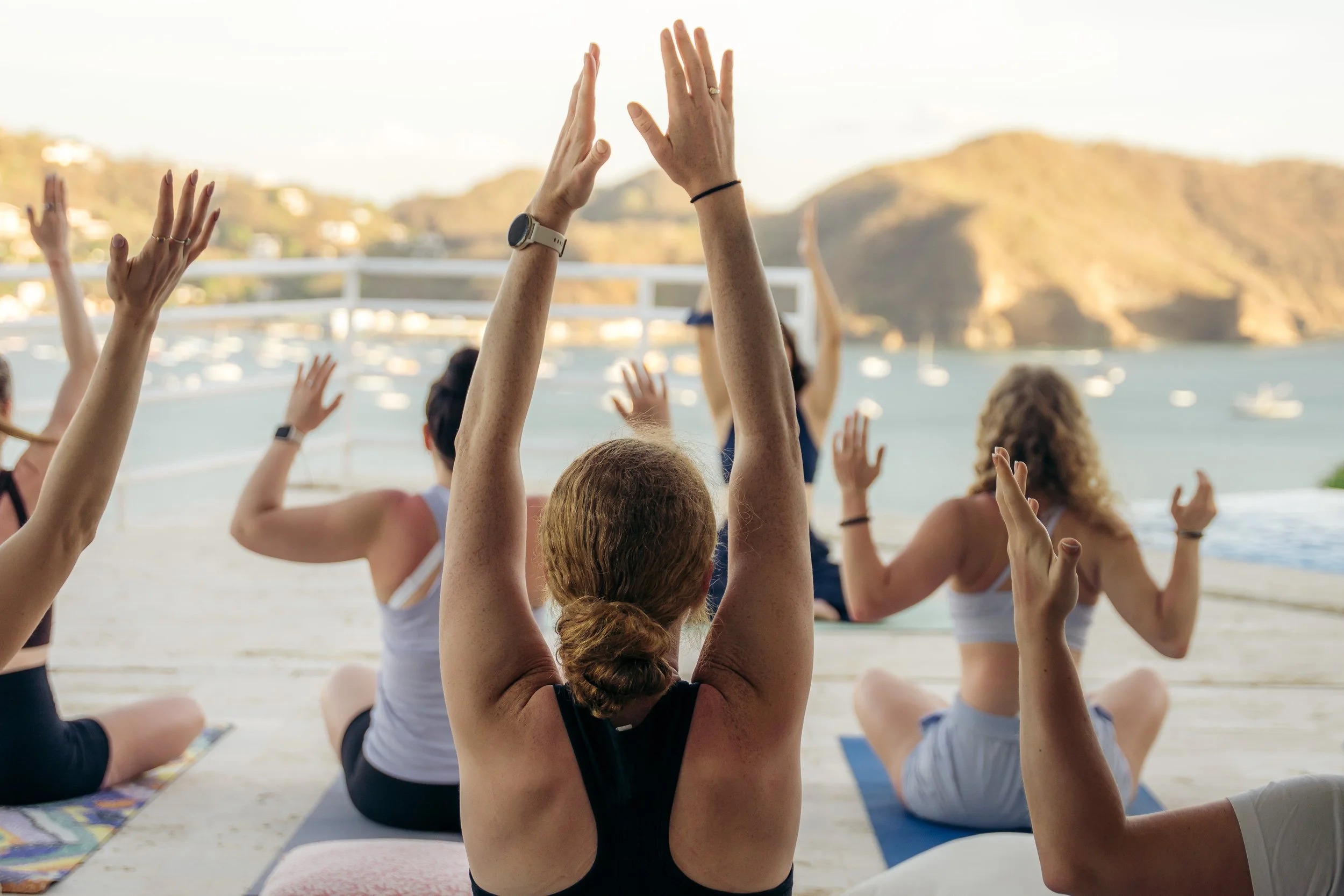 People participating in a yoga class outdoors on a deck overlooking the water and hills, sitting cross-legged with arms raised in a yoga pose.