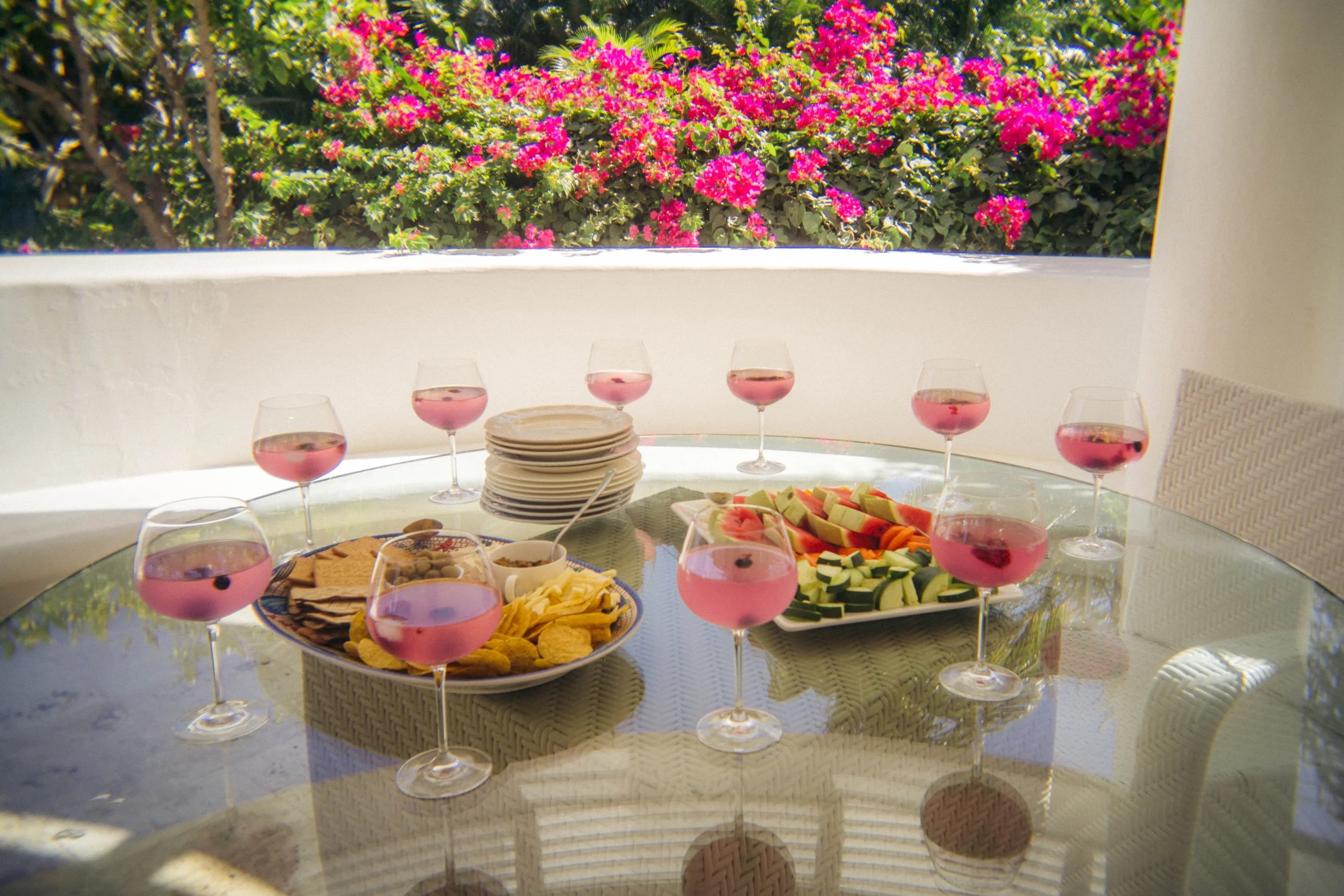A glass table set with a platter of crackers, a bowl of snacks, and a plate of sliced fruits consisting of watermelon, cantaloupe, and zucchini, with seven glasses of pink beverage on an outdoor patio. Pink flowering plants are visible in the background.