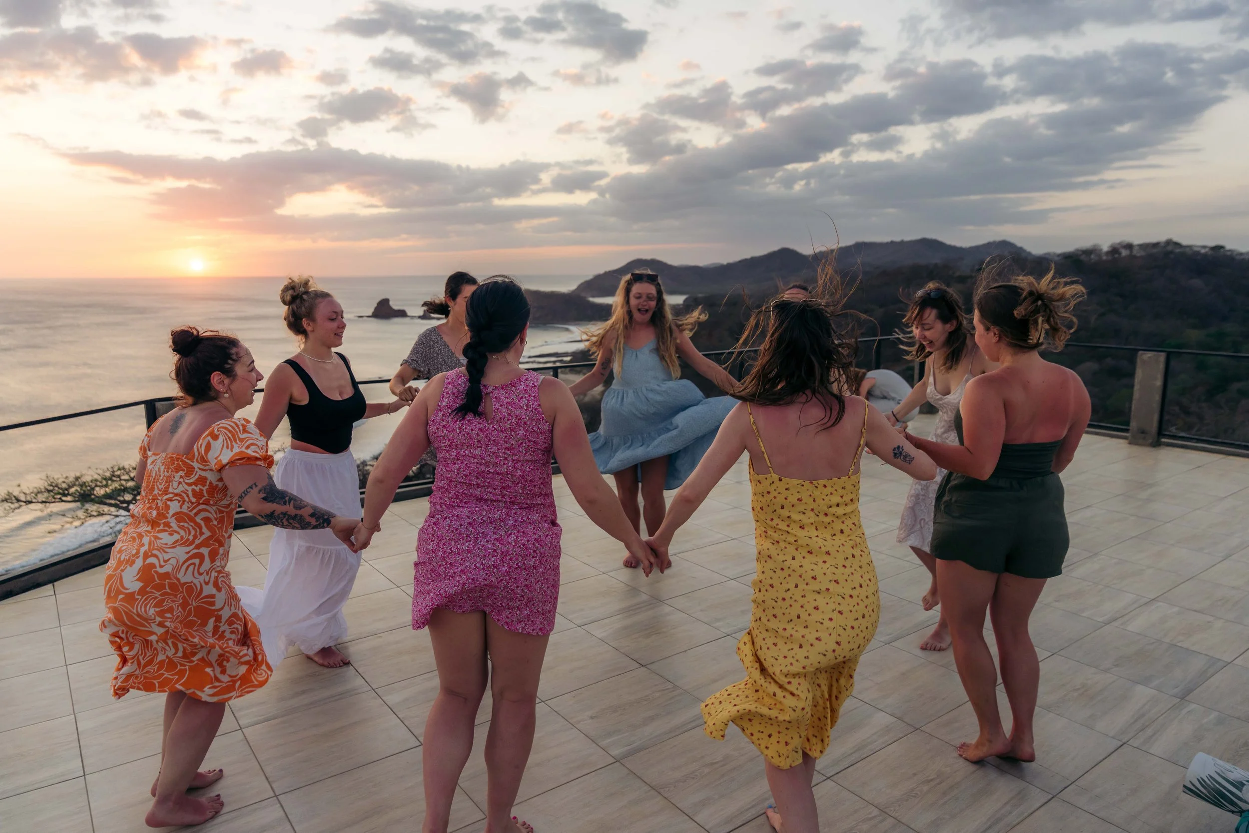 A group of women holding hands and dancing in a circle on a rooftop at sunset with ocean and hills in the background.