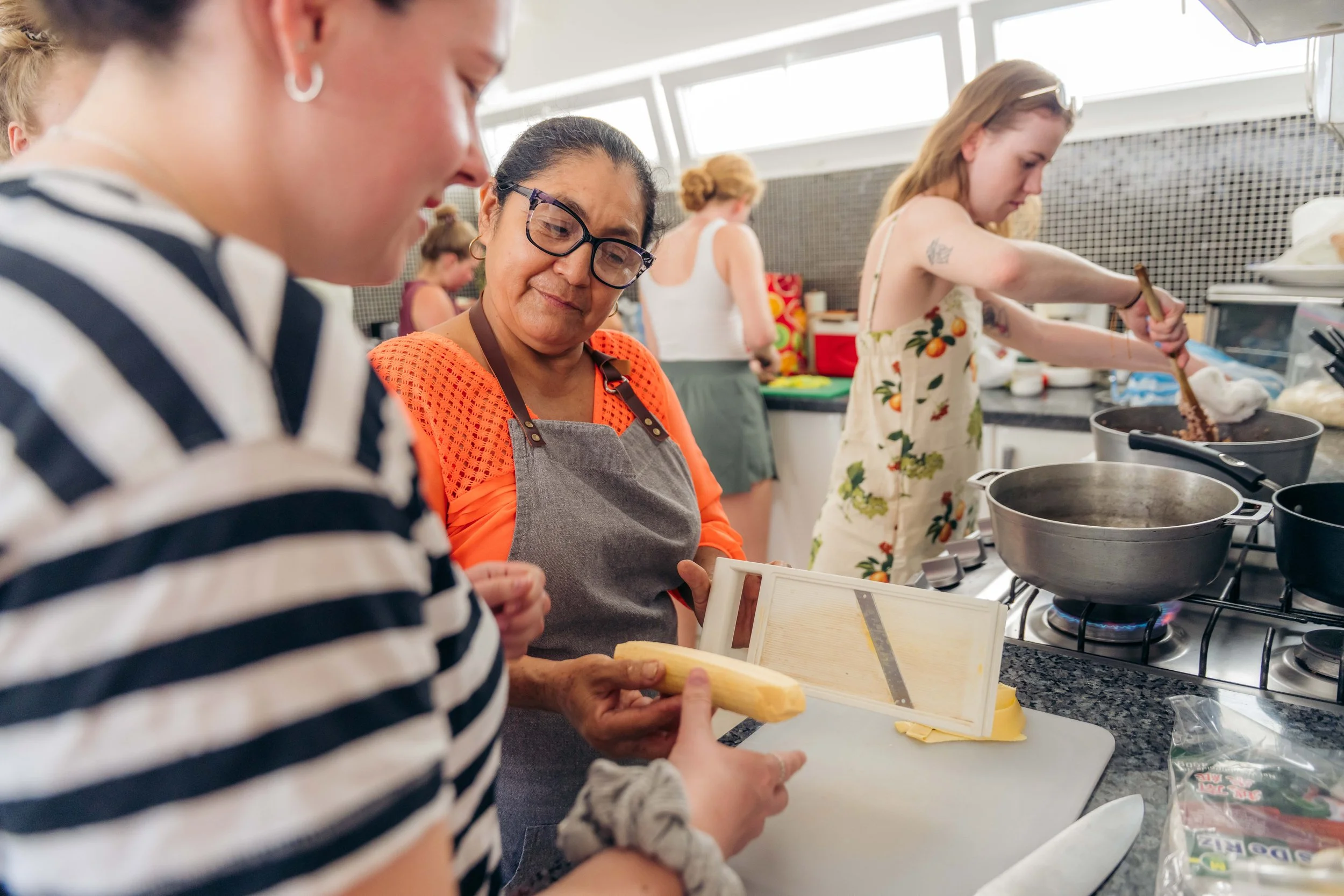 Multiple women are cooking in a kitchen with gas stoves. They are preparing food and using various kitchen tools and ingredients.