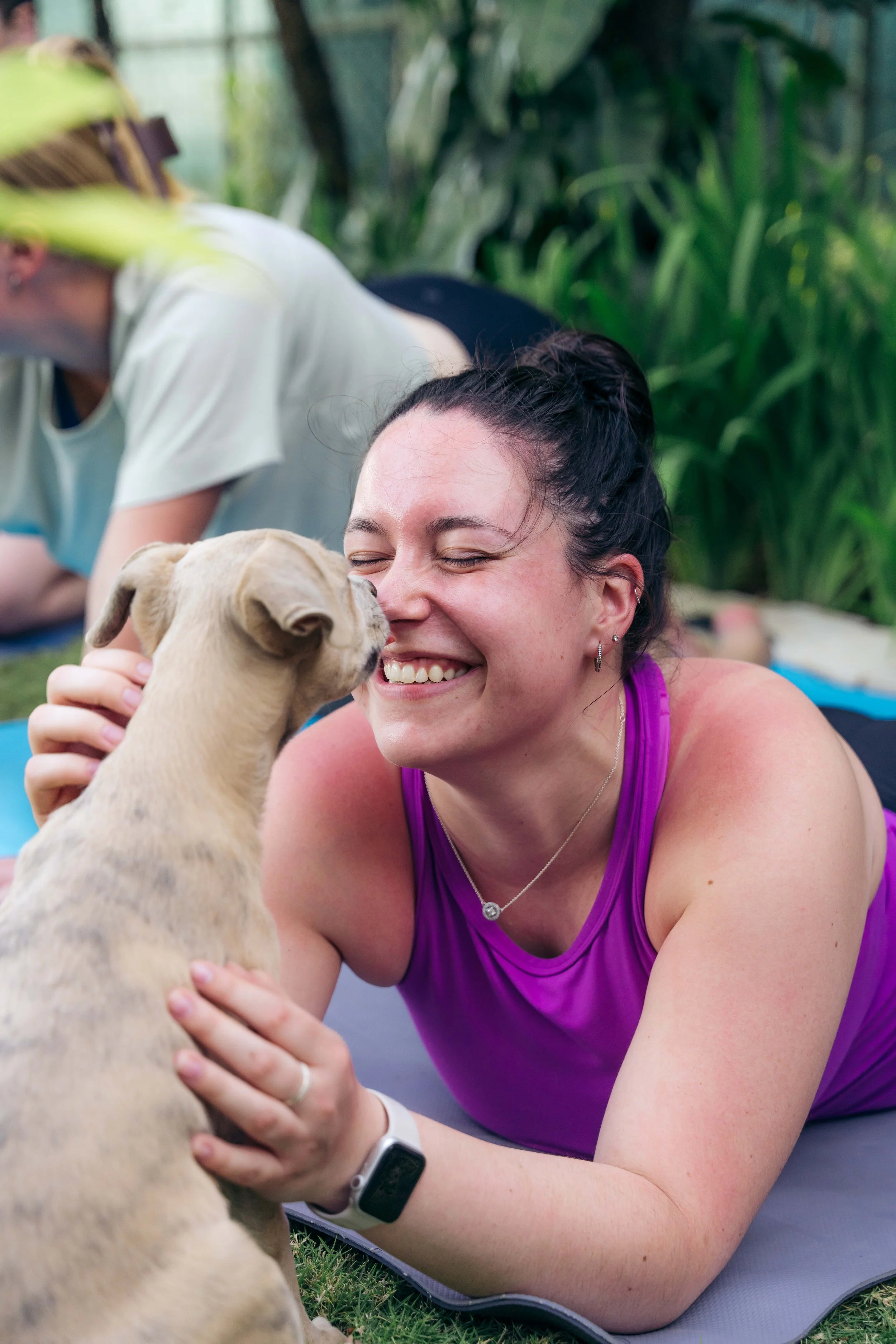 A smiling woman lying on a yoga mat outdoors, touching a puppy's face, with green plants in the background.