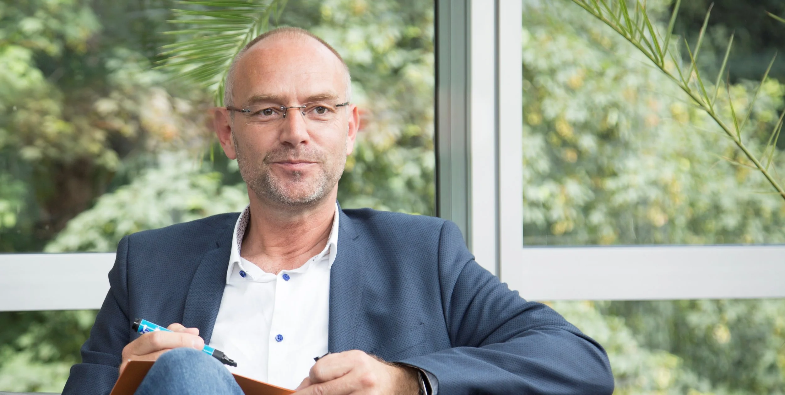 Frau sitzt erschöpft am Tisch mit Laptop, Kaffee und Notizbuch, reibt sich die Augen. Sie trägt eine Brille und ein rot gemustertes Oberteil.