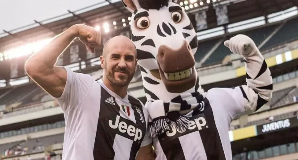 A soccer player, in a Juventus jersey, standing next to a person in a zebra mascot costume, both flexing their arm muscles in a stadium.