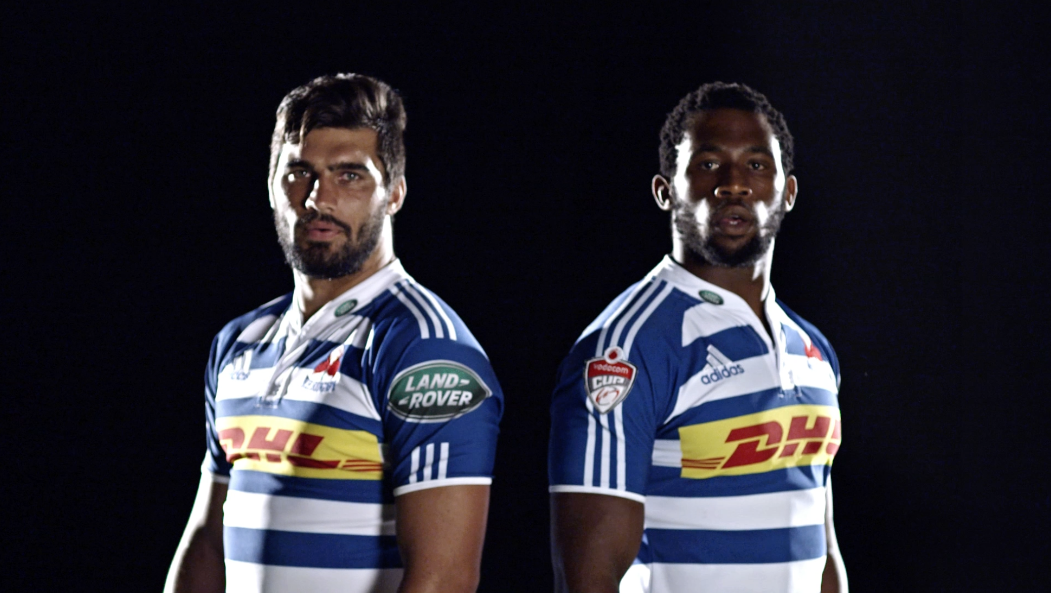 Two rugby players in blue and white striped jerseys standing back to back against a black background.